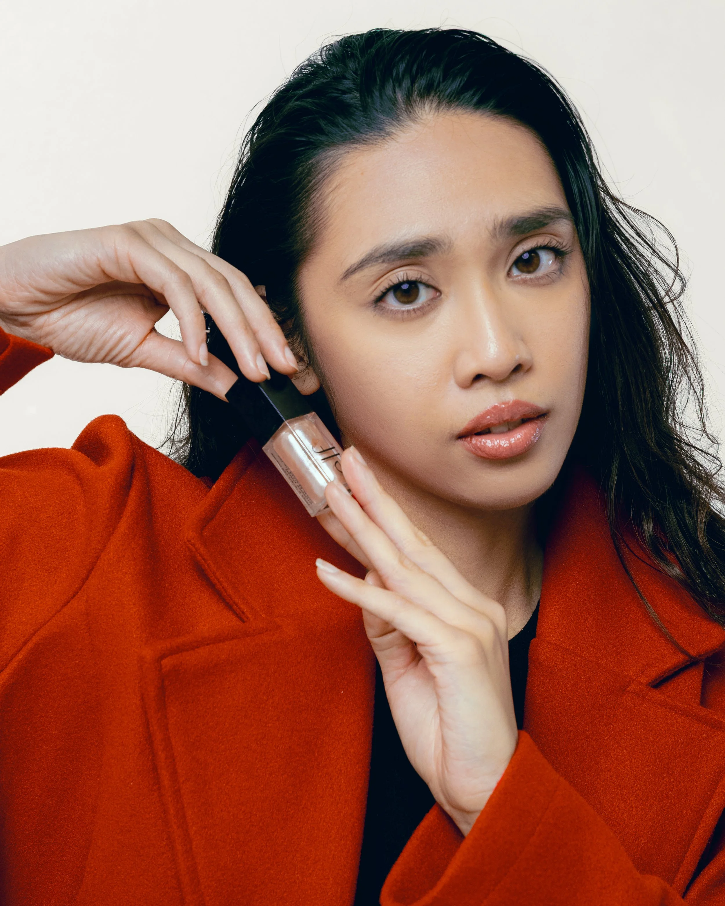 A young woman with long dark hair holding a small bottle of nail polish near her face, wearing a red coat.