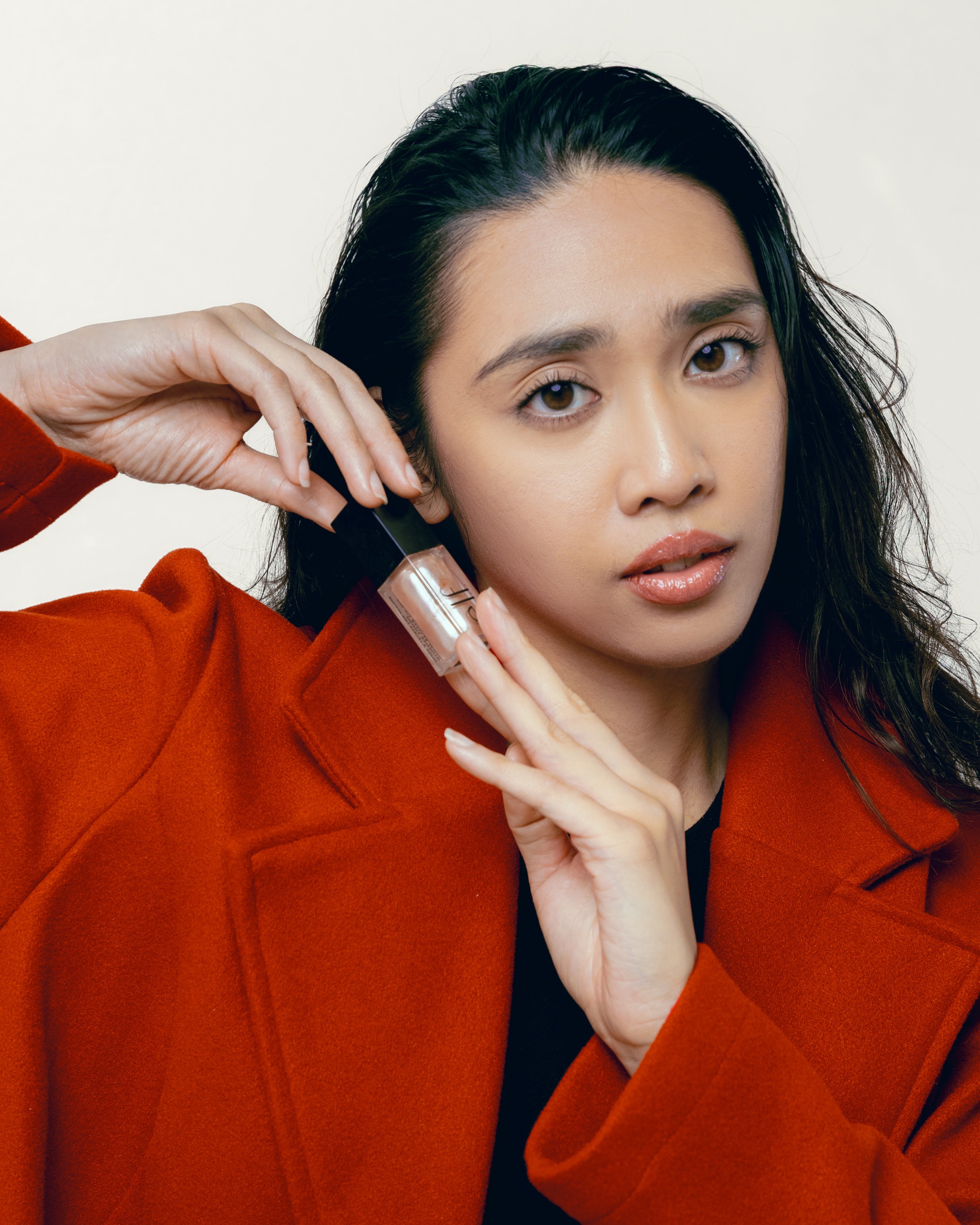 A woman with long dark hair is holding a small bottle of nude nail polish near her face, wearing an orange coat and a black top, against a plain white background.