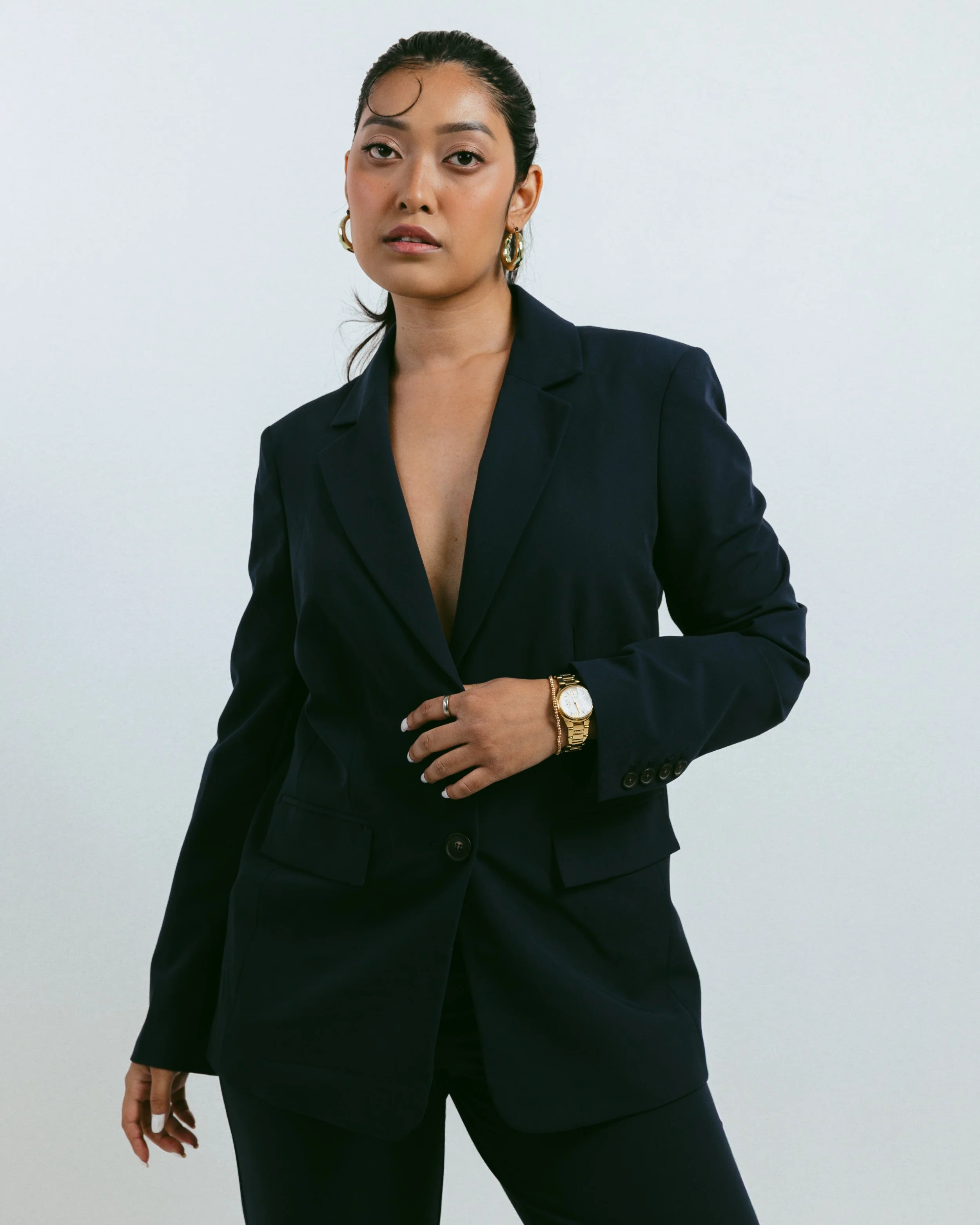 A woman in a black suit with gold jewelry and a watch, standing against a plain white background.