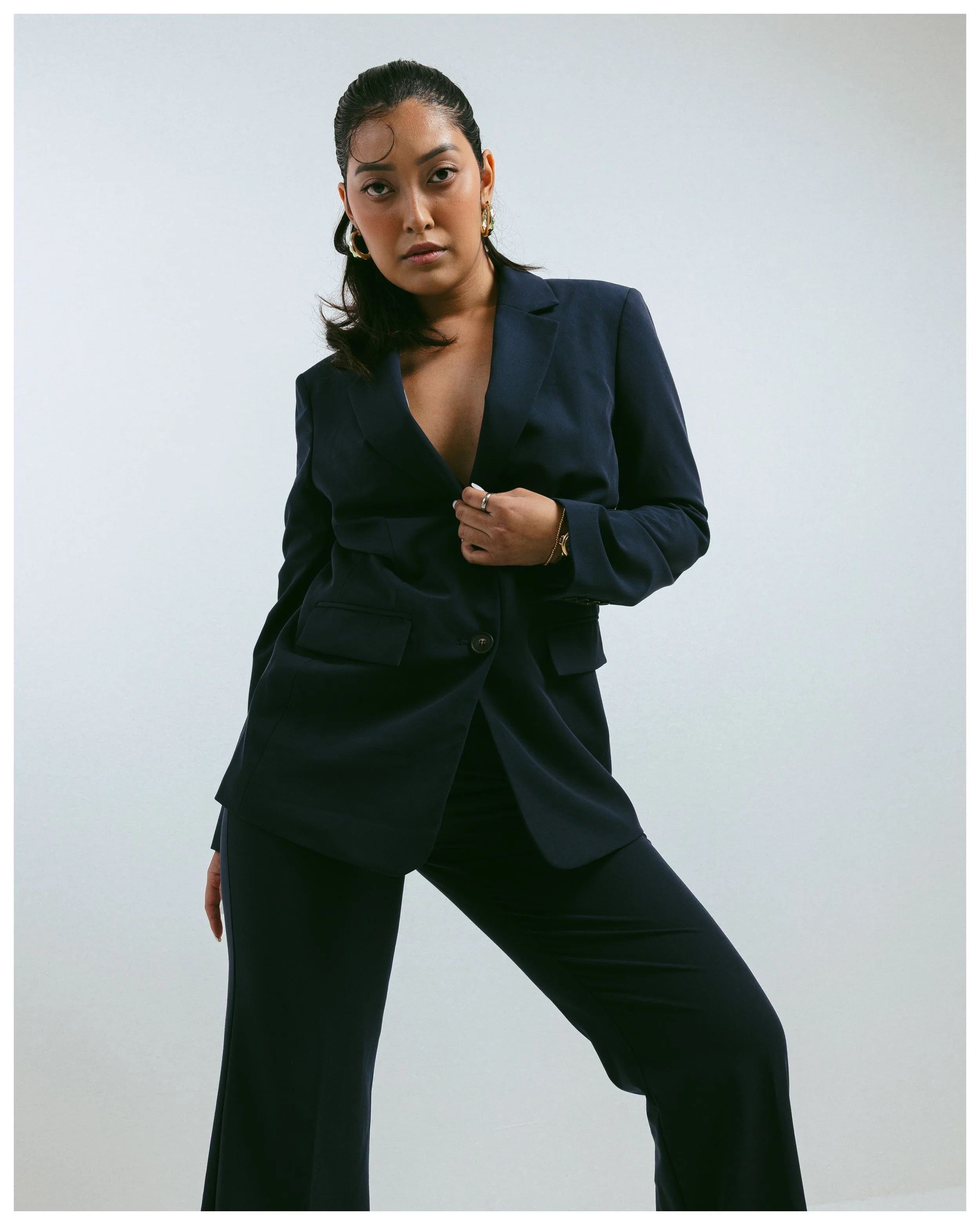 A woman dressed in a dark blazer and pants, posing against a plain light background. She accessorizes with gold hoop earrings, a bracelet, and a ring, with one hand on her chest and the other hanging down.