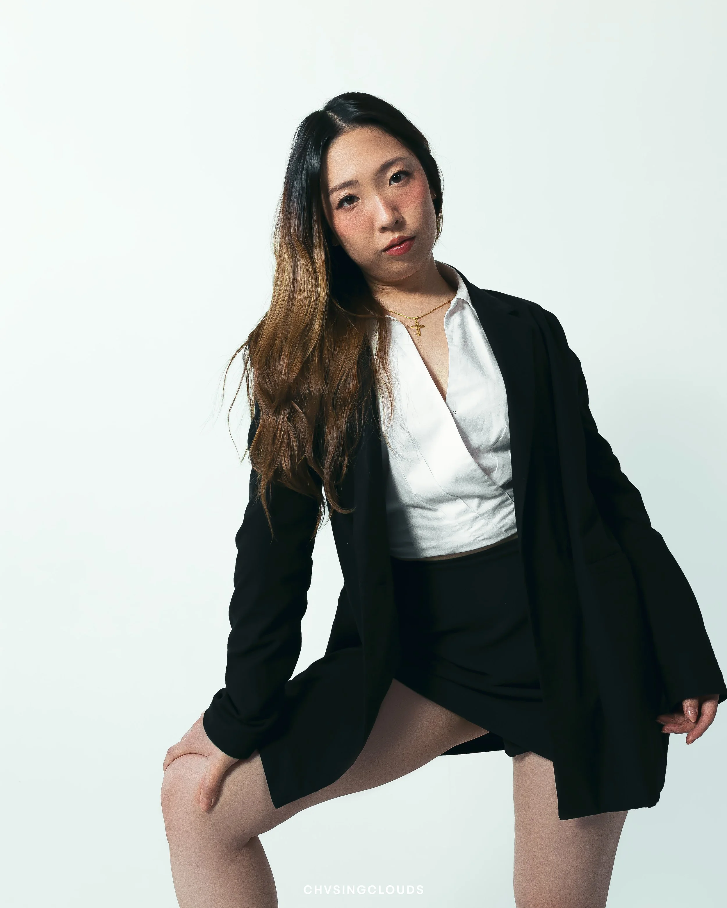 Young woman with long wavy hair, wearing a black blazer, white shirt, black skirt, and ginger tights, posing with one hand on her knee in front of a plain light background.