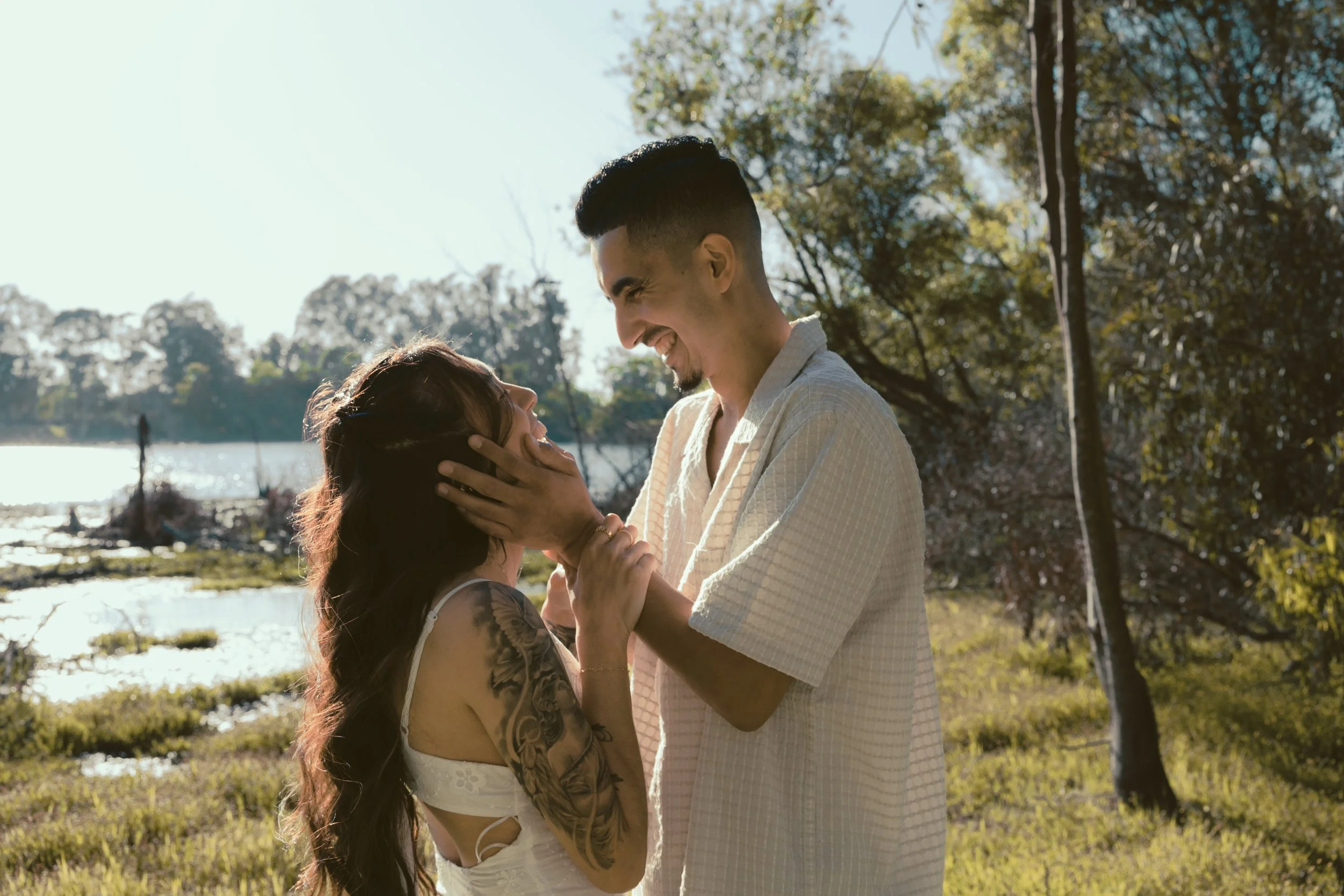 A couple standing outdoors by a river, smiling and looking at each other, with trees and sunlight in the background.