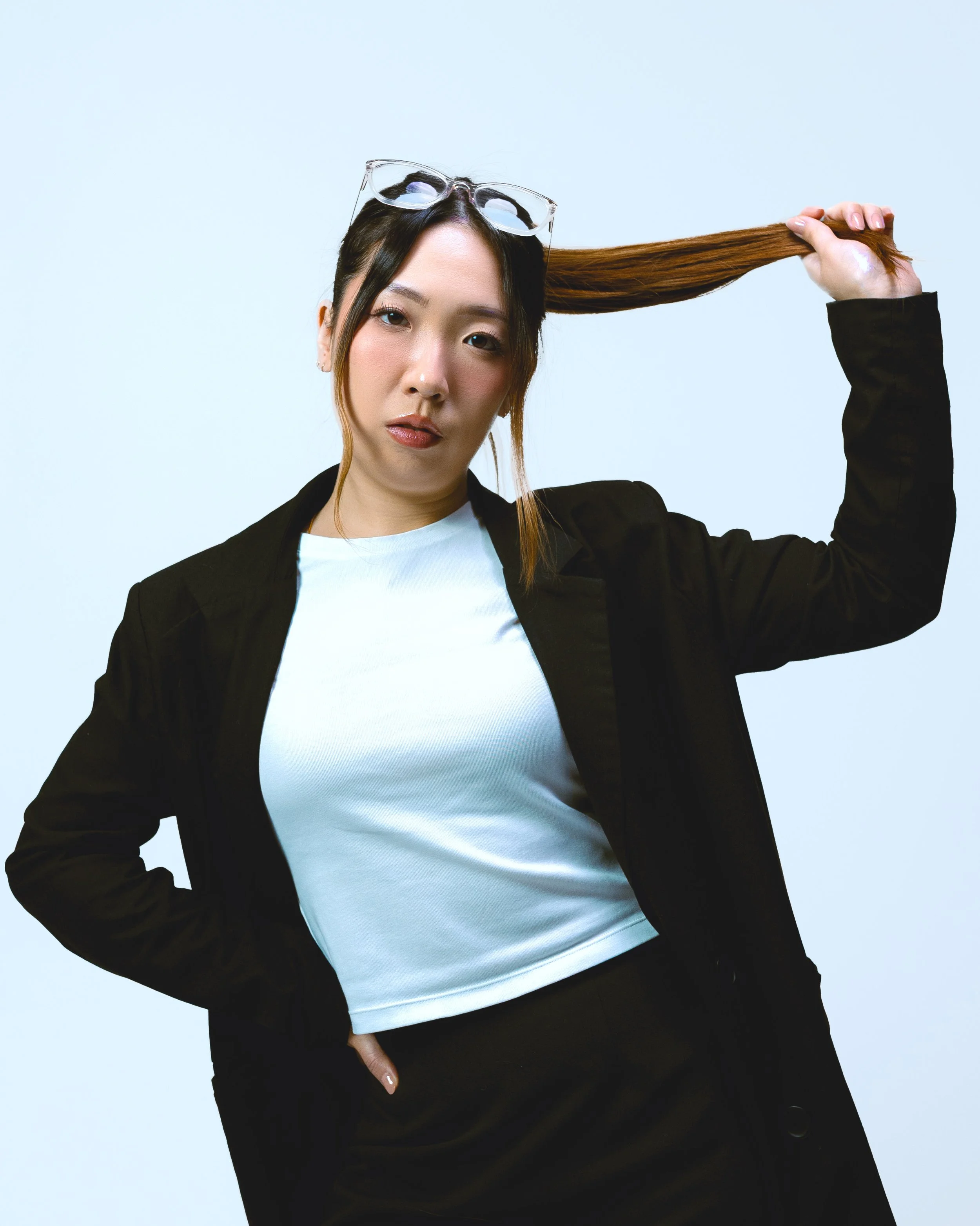 A woman with glasses on her head, wearing a black jacket and white shirt, holds her long brown hair with one hand while looking at the camera.