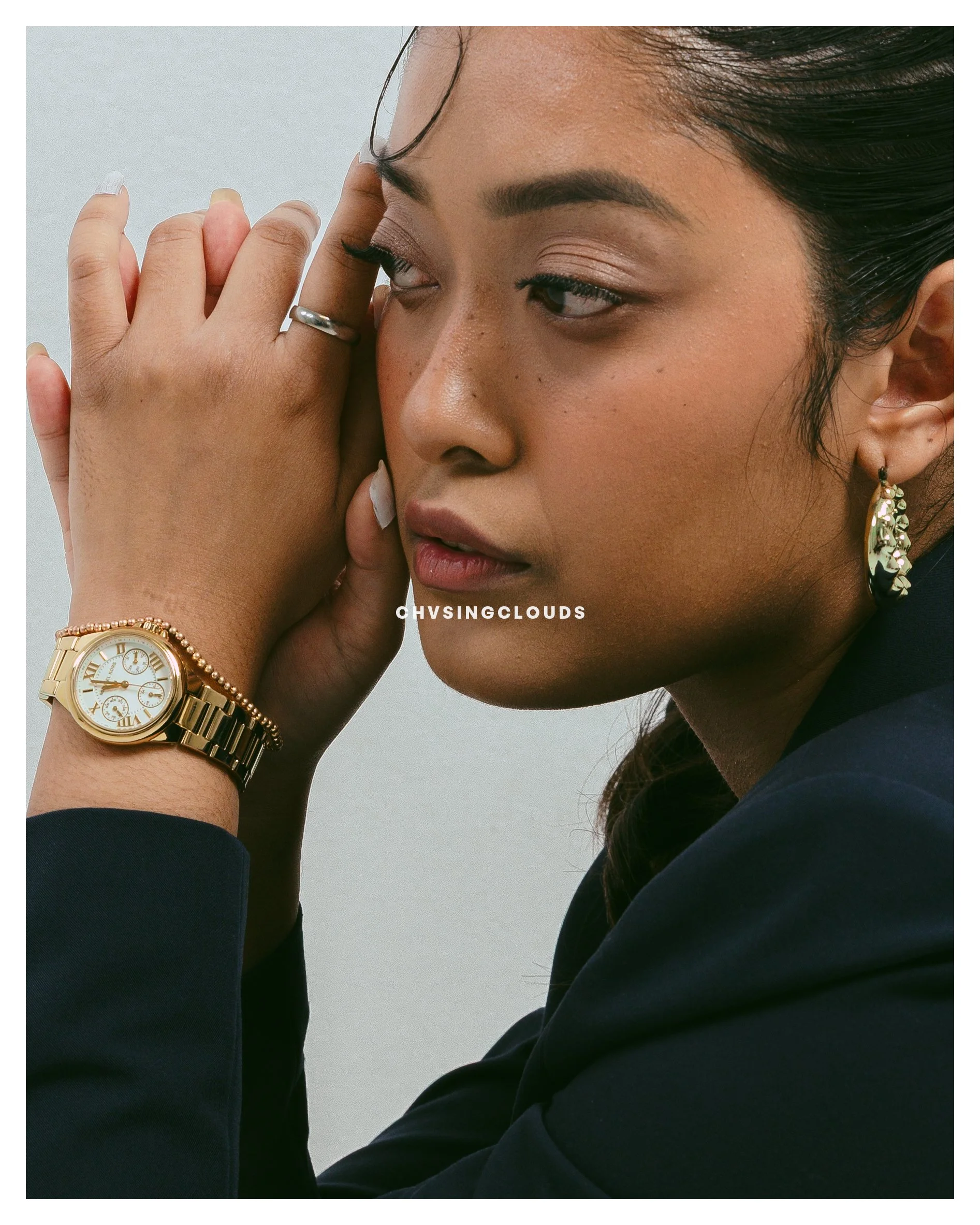 Close-up of a woman with dark hair wearing makeup, gold earrings, a gold watch, and a ring, touching her temple with her hand.