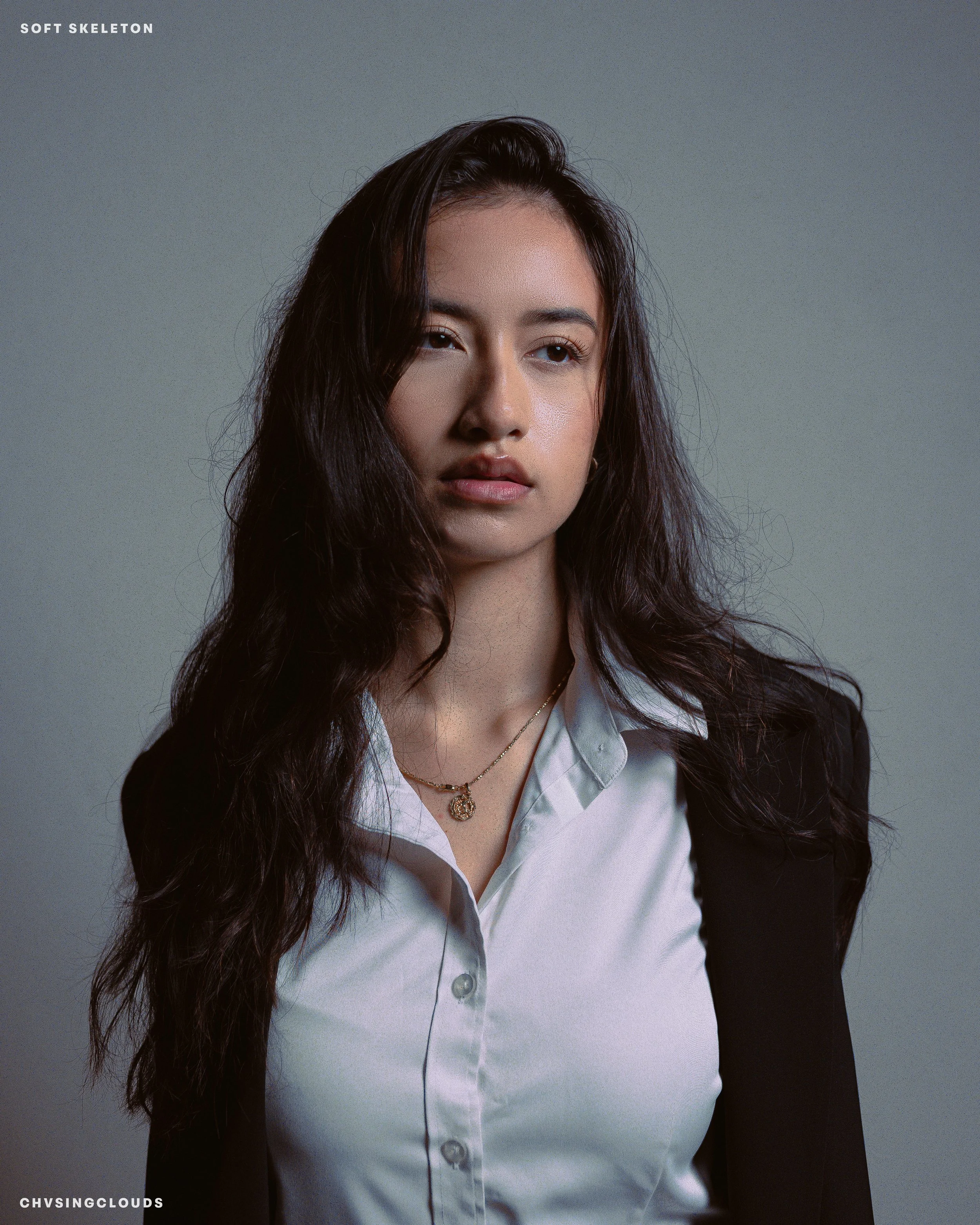 A young woman with long, wavy dark hair, wearing a white collared shirt and a black blazer, looking slightly to the side with a neutral expression. She is also wearing a gold necklace with a pendant.