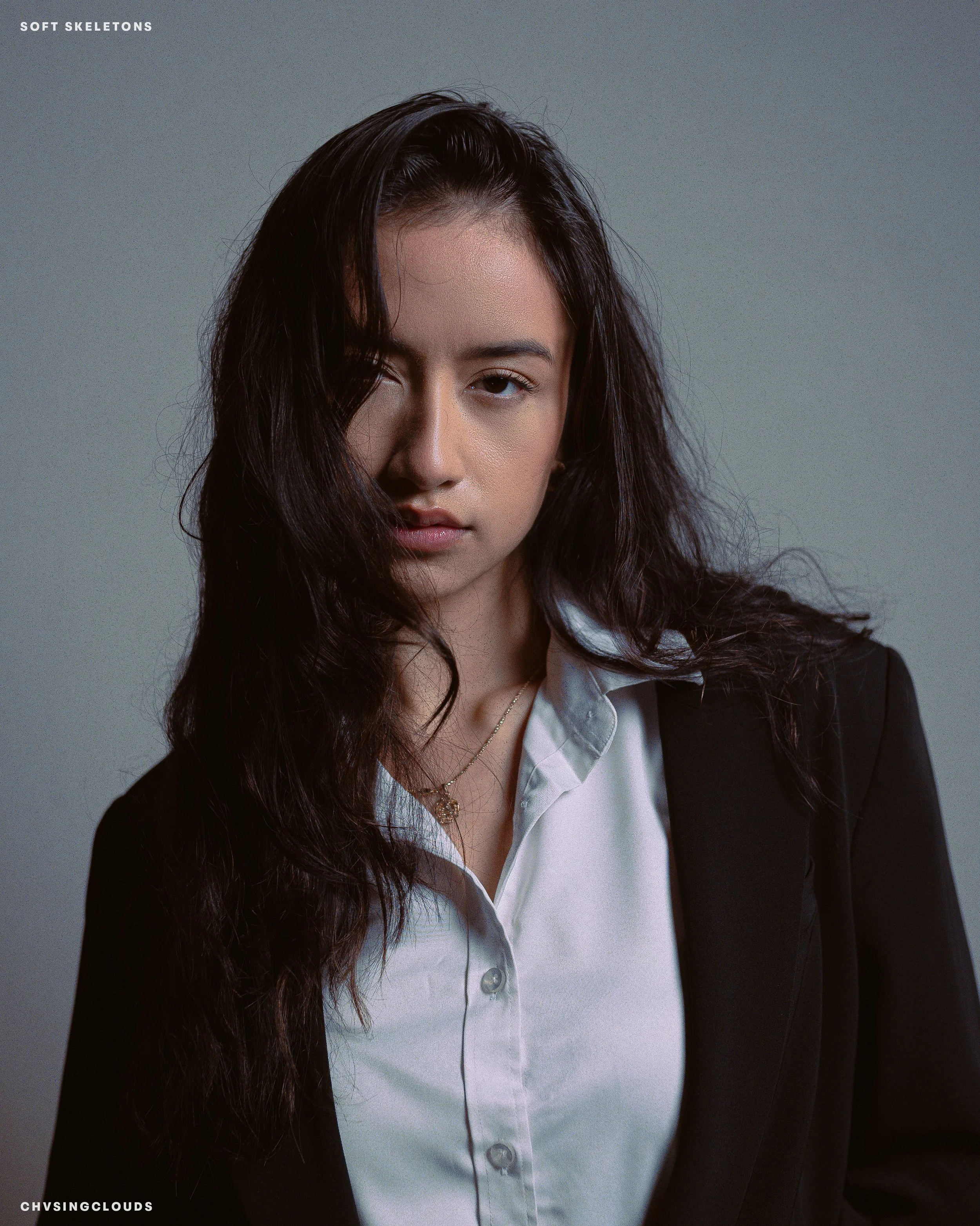 A woman with wavy dark hair, wearing a white blouse and black blazer, looking directly at the camera with a serious expression.