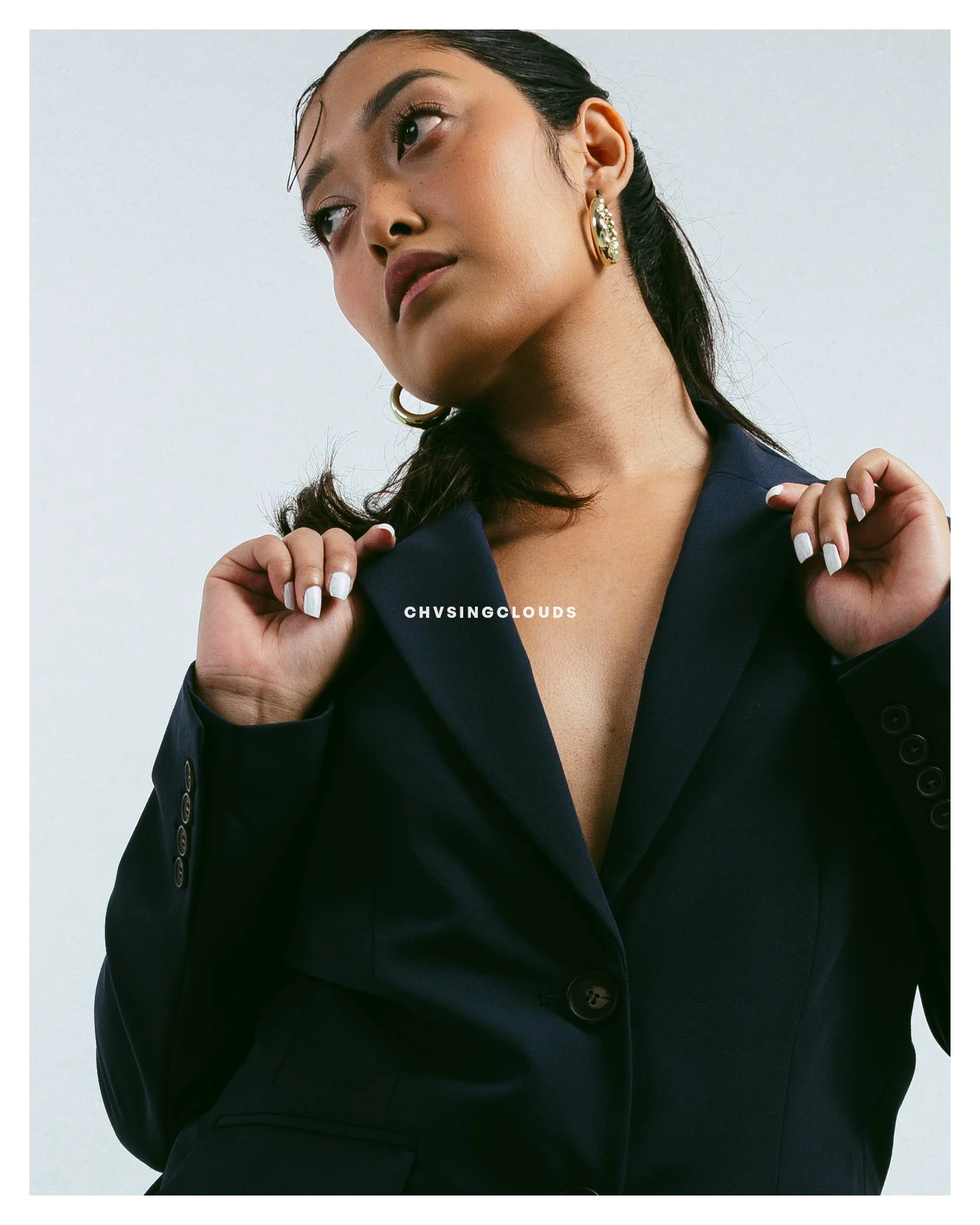 A woman with dark hair, wearing a navy blazer, holding the lapels, and showing her neck and earrings against a plain light background.