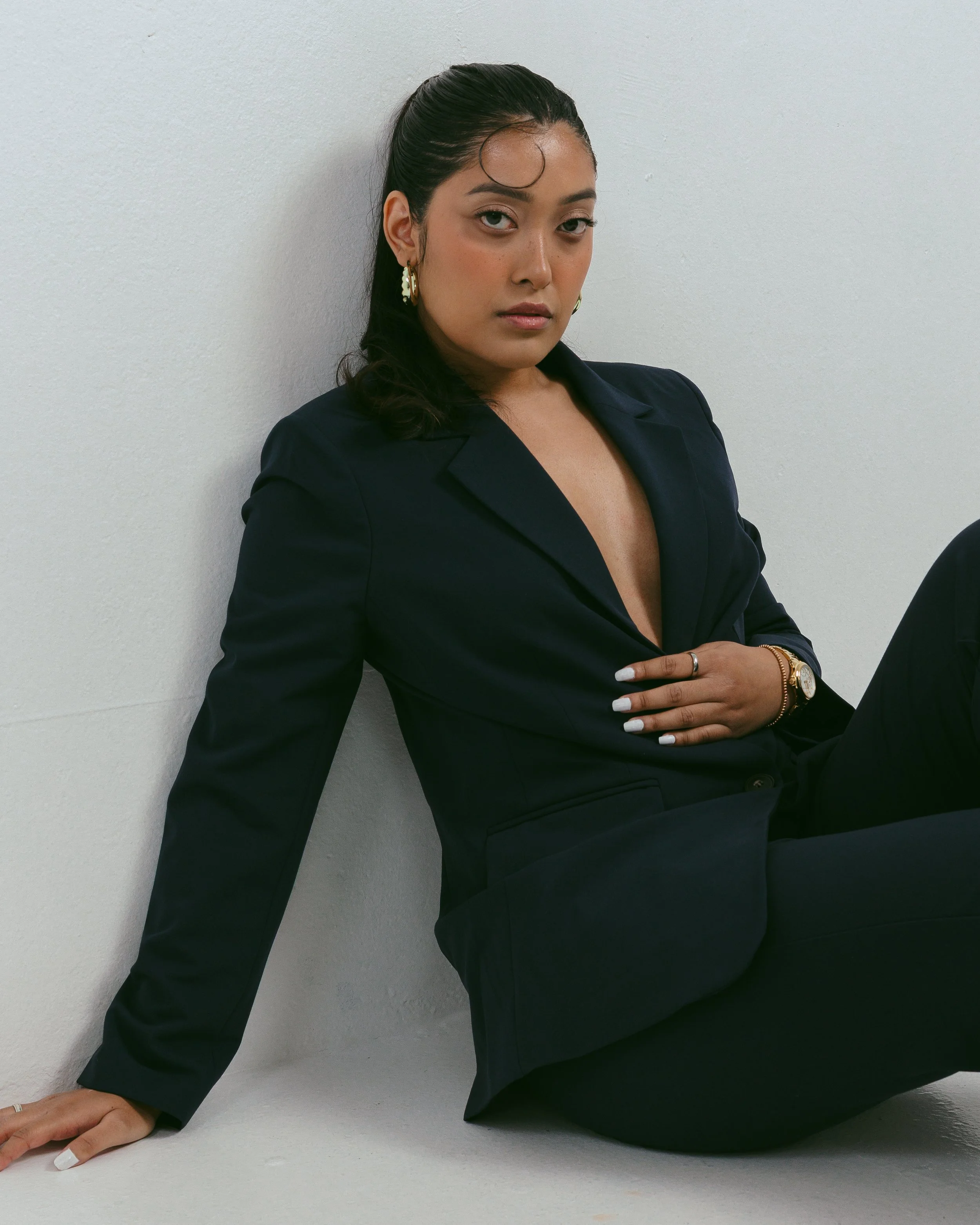 Woman in a dark blazer sitting on the floor, leaning against a white wall, with one arm extended and hand resting on the floor, wearing gold earrings and a watch.