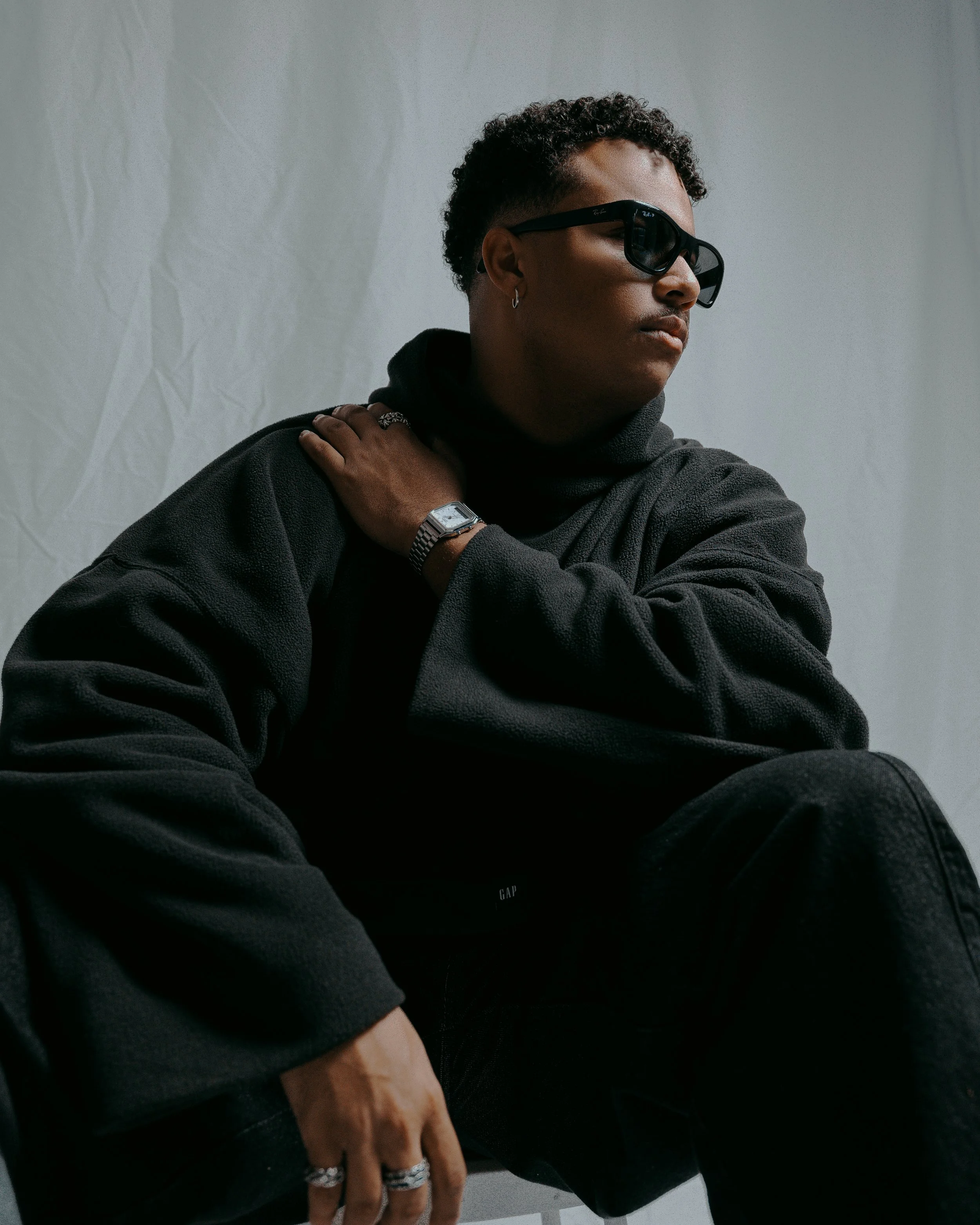A person with short curly hair, wearing sunglasses, a black hoodie, and black pants, sitting against a white background, and looking to the side.