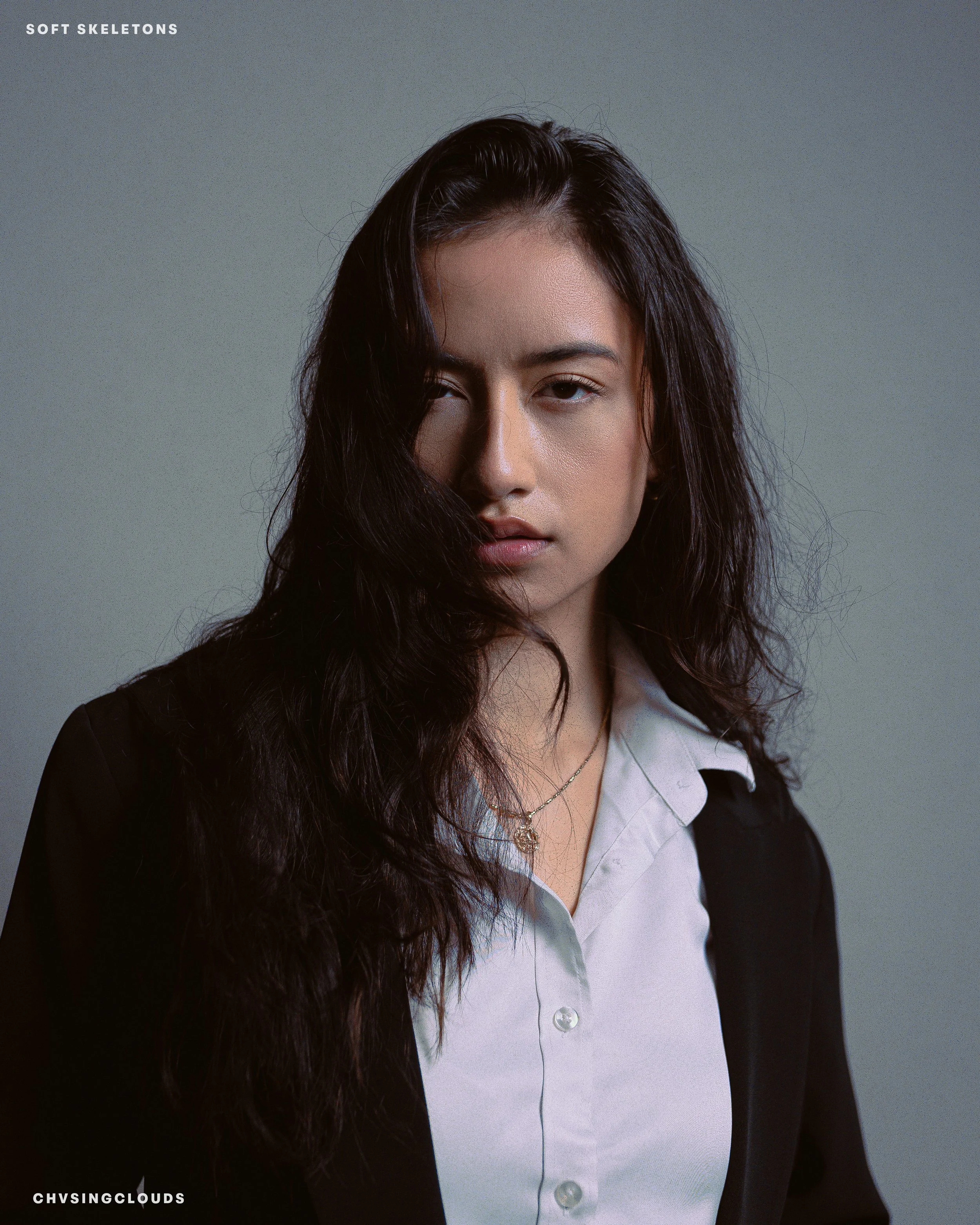 Portrait of a young woman with wavy dark hair, partially covering her face, wearing a white shirt and a black blazer against a plain gray background.