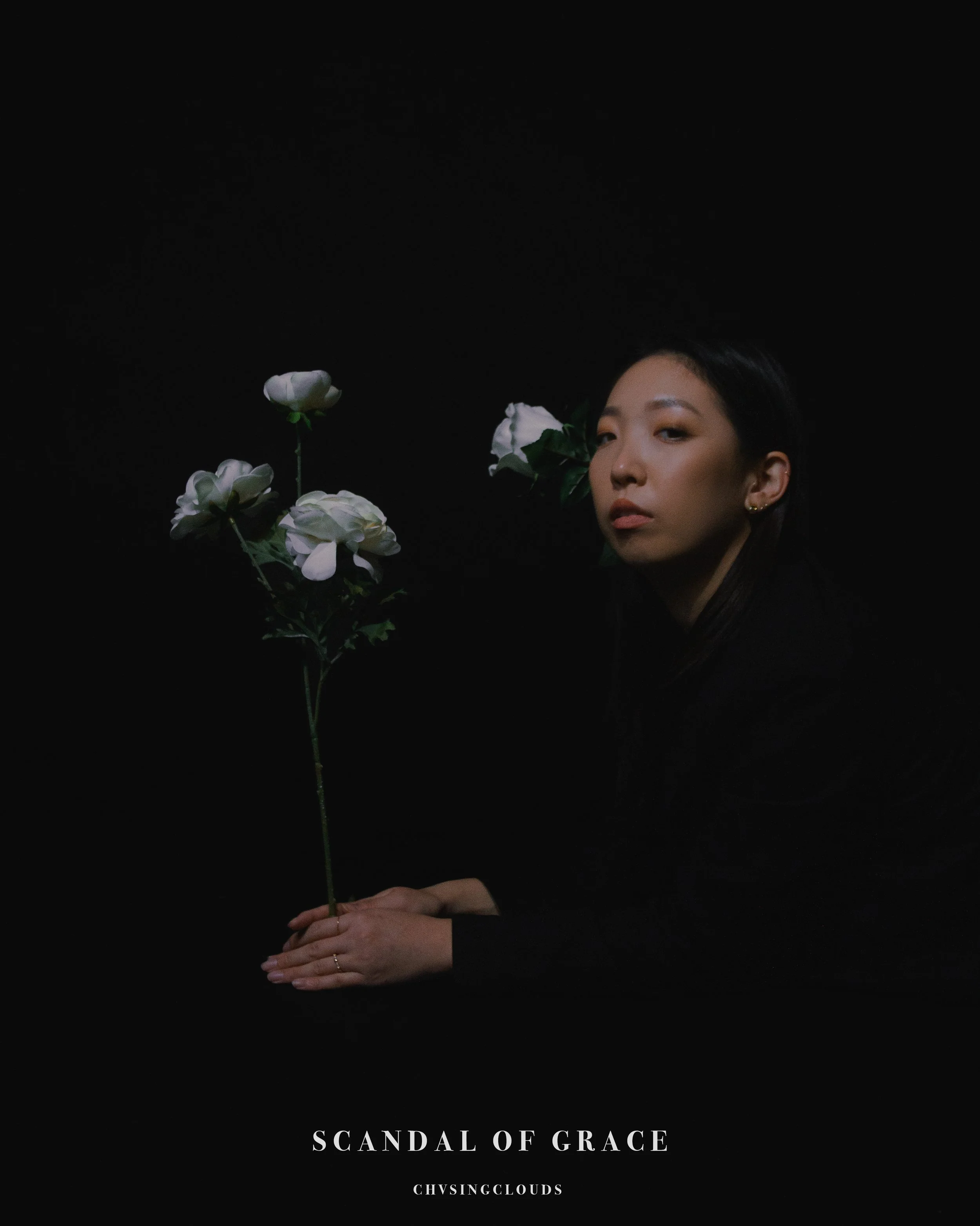 A woman with dark hair and earrings holding a white flower against a black background, with the caption 'Scandal of Grace' and the handle '@chvsingclouds'.