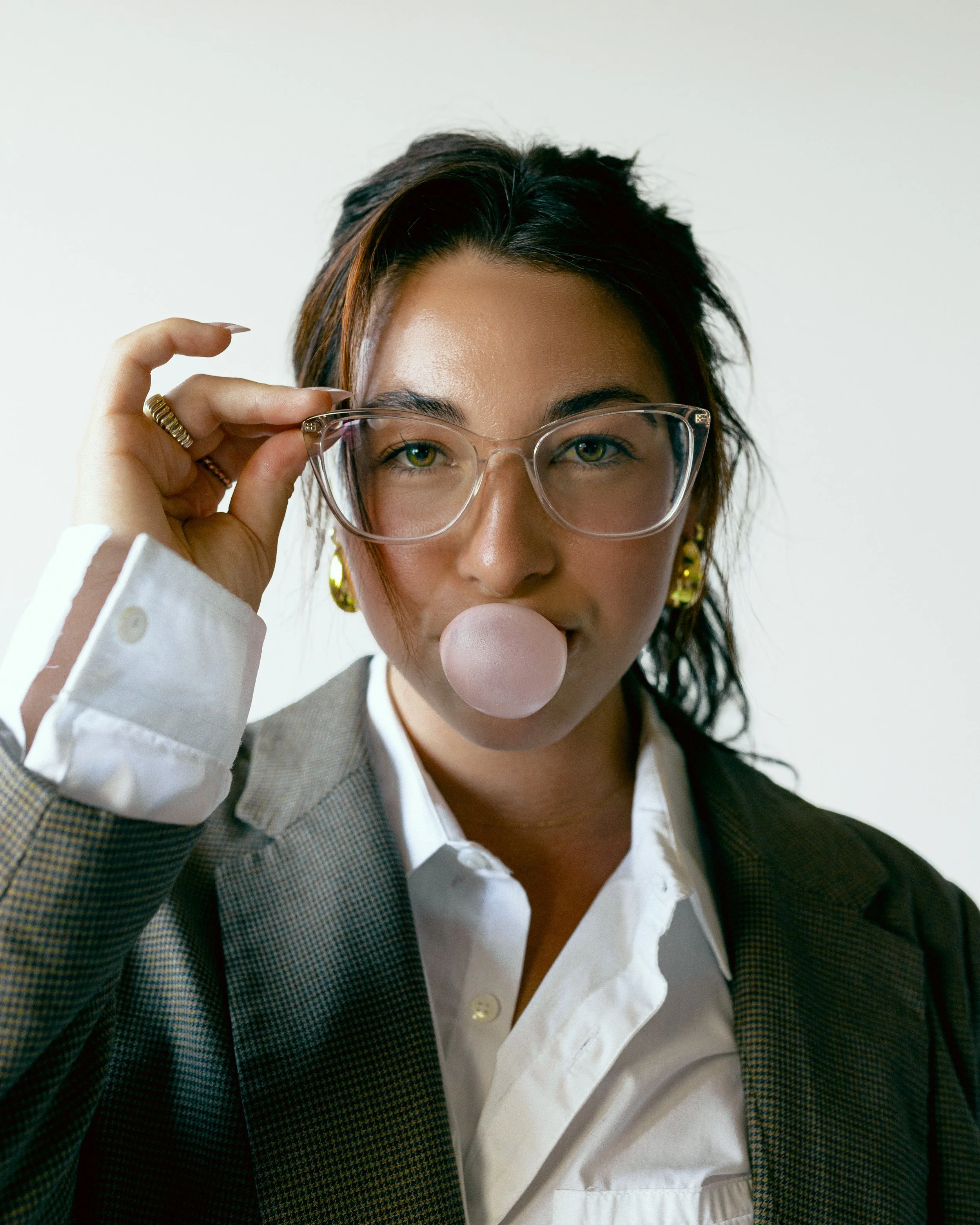 A woman with dark hair, glasses, and gold earrings blows a bubble with bubble gum, while wearing a white shirt and a plaid blazer.