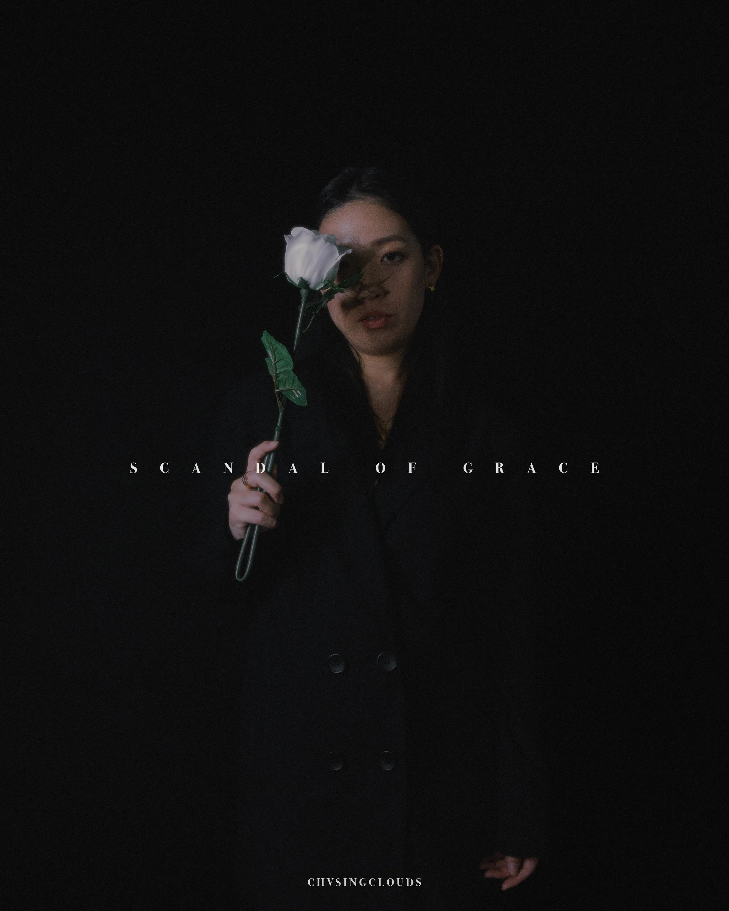 A woman in dark clothing holding a white rose in front of her face, partly covering one eye, against a black background, with the words 'Scandal of Grace' across her chest.