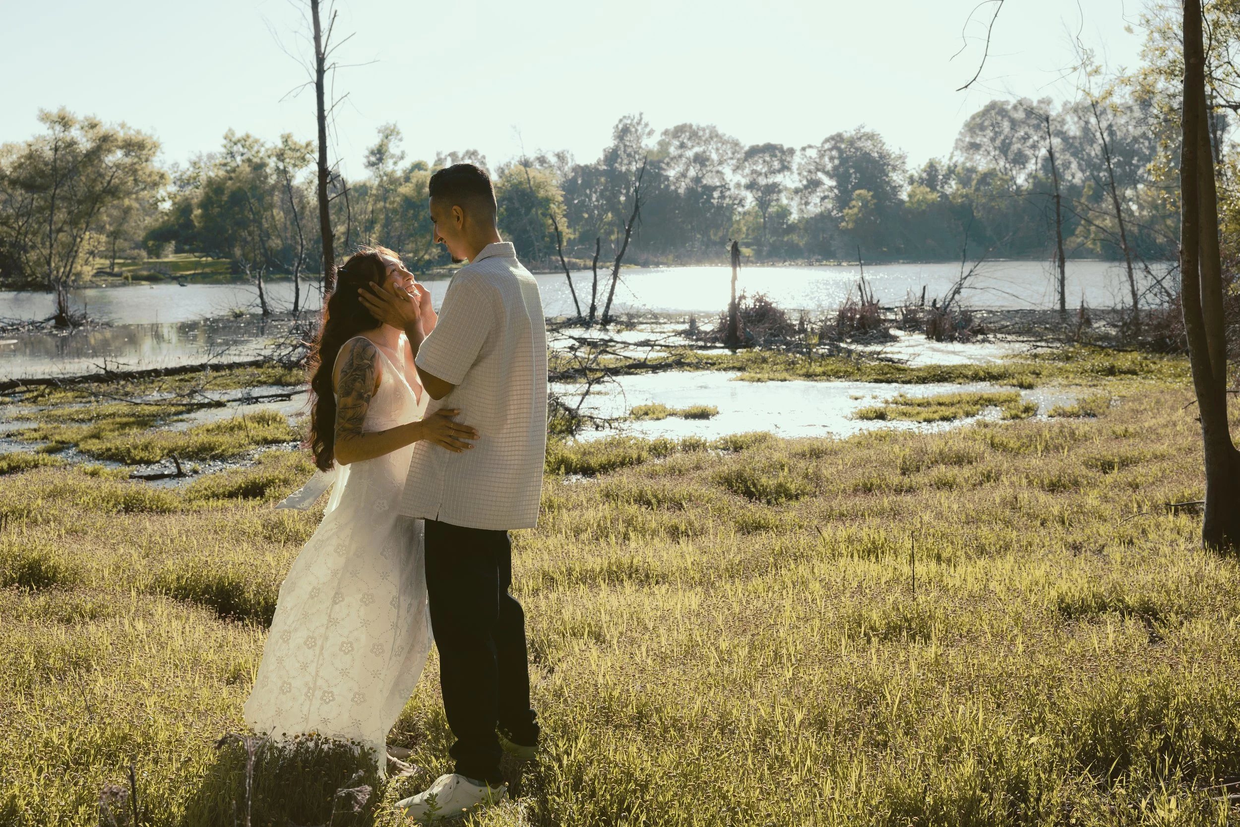 A couple in wedding attire standing in a grassy area near a body of water, smiling and looking at each other, with trees and sunlight in the background.