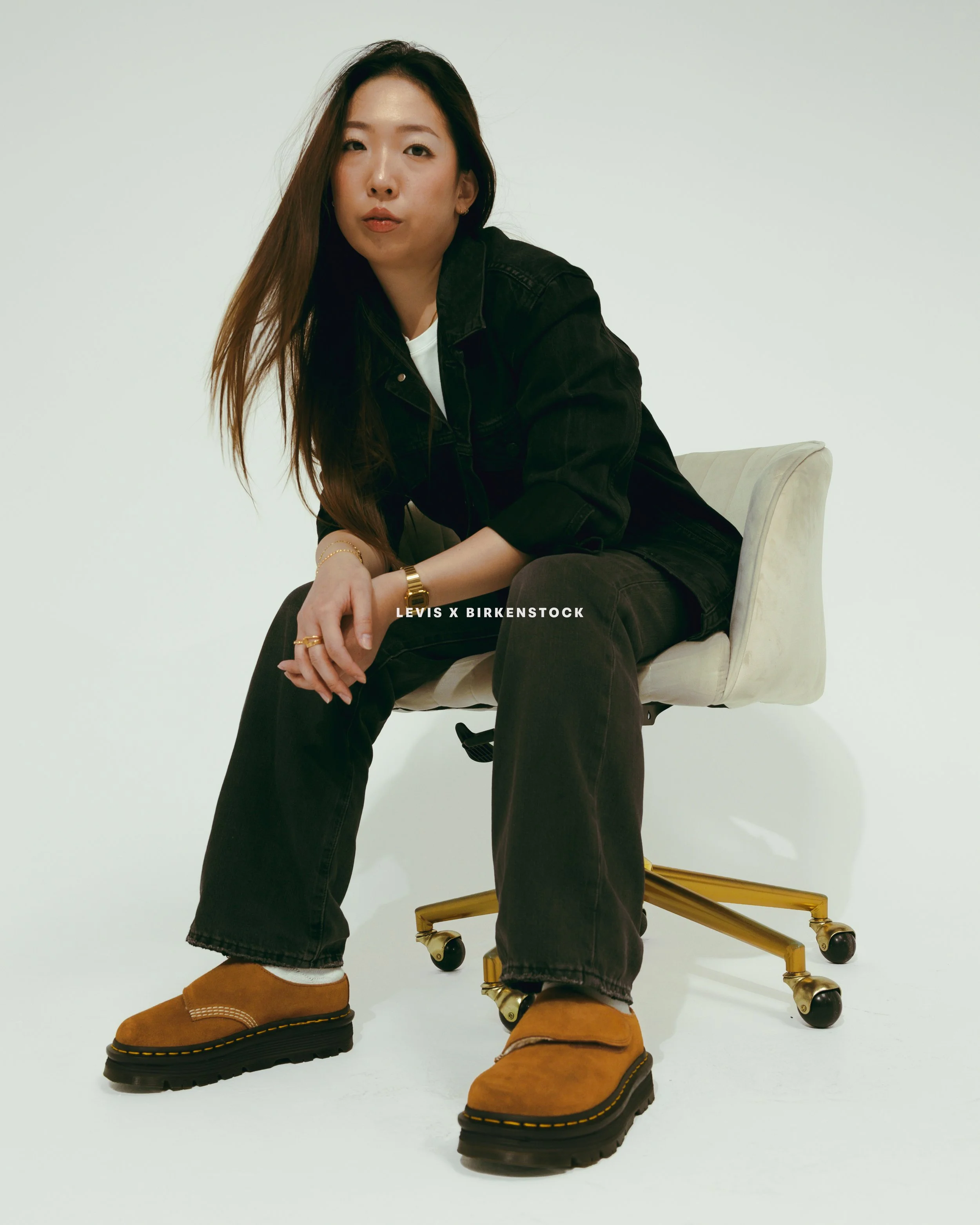 Person sitting on a white chair, wearing tan boots, black jeans, a white shirt, and a black jacket, with a white background.