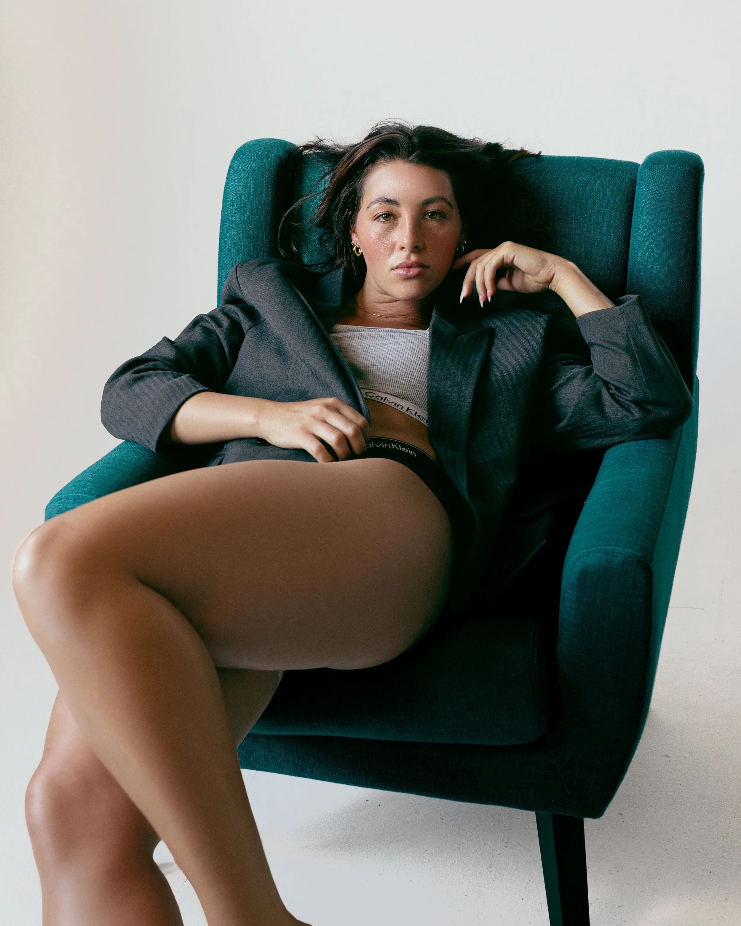 A woman sitting comfortably in a teal armchair, wearing a black blazer, Calvin Klein underwear, and gray top, with one leg crossed over the other, looking directly at the camera with a serious expression.