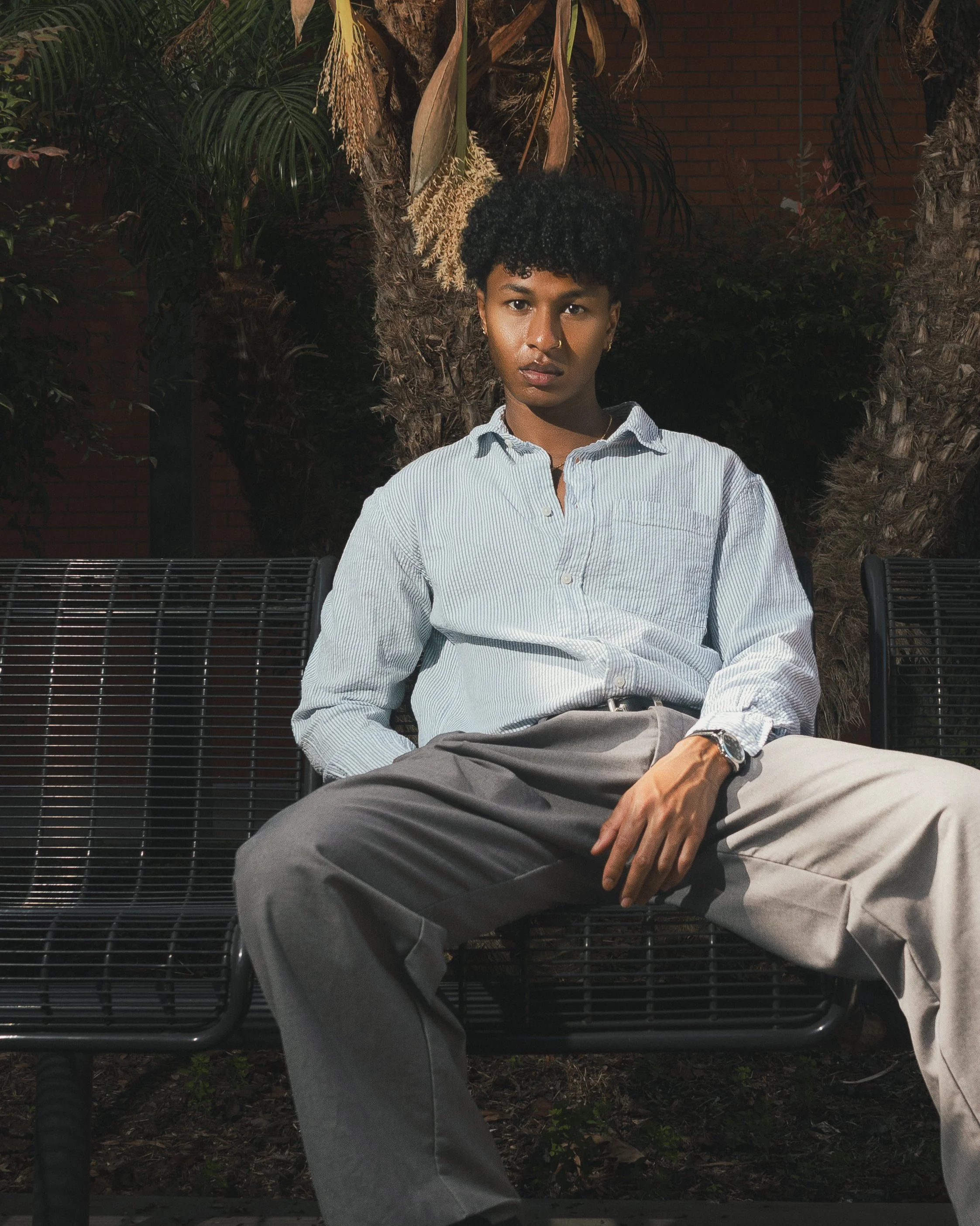 A young person with curly hair sits on a black metal bench outdoors at night, wearing a light striped shirt and beige pants, with palm trees and a brick wall in the background.