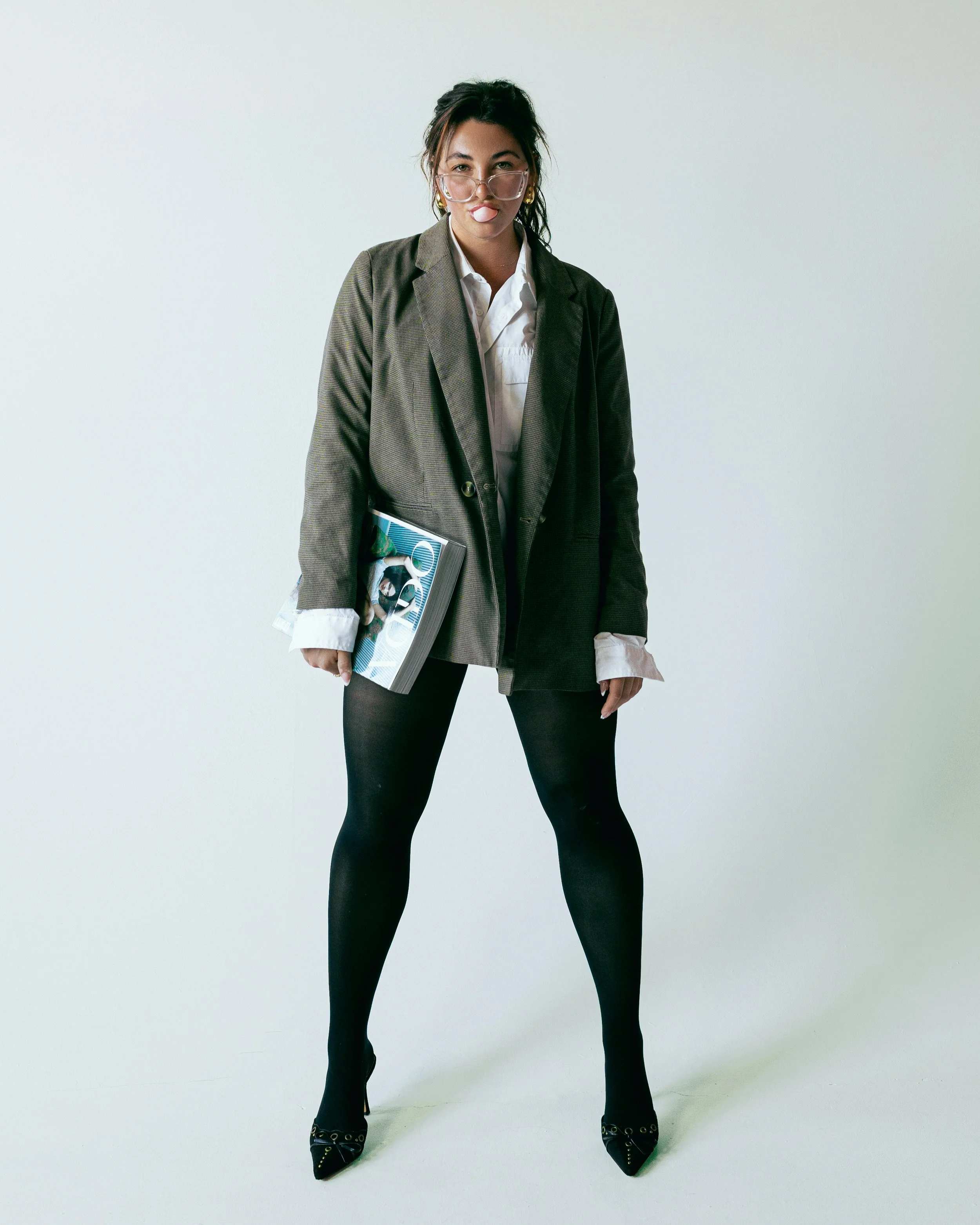 A woman standing against a plain light-colored background, wearing a blazer, white shirt, black tights, and high heels, holding a magazine or notebook, with glasses and a lip balm or similar item in her mouth.
