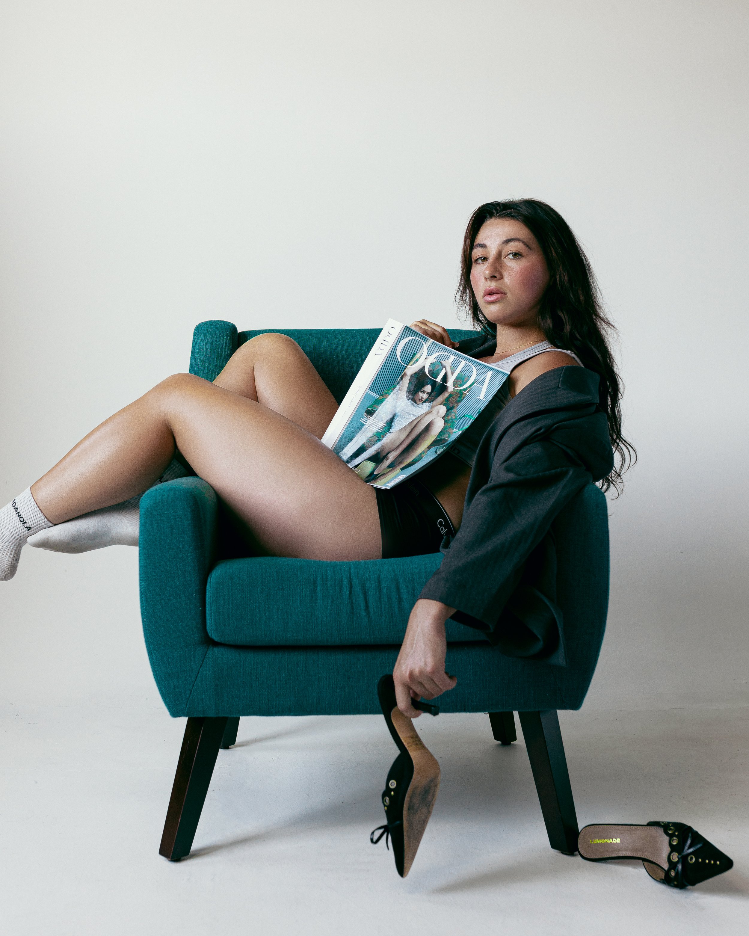 Young woman sitting in a teal armchair with removed black high heels, holding a Vogue magazine, wearing a gray sleeveless top, black shorts, and a black blazer.