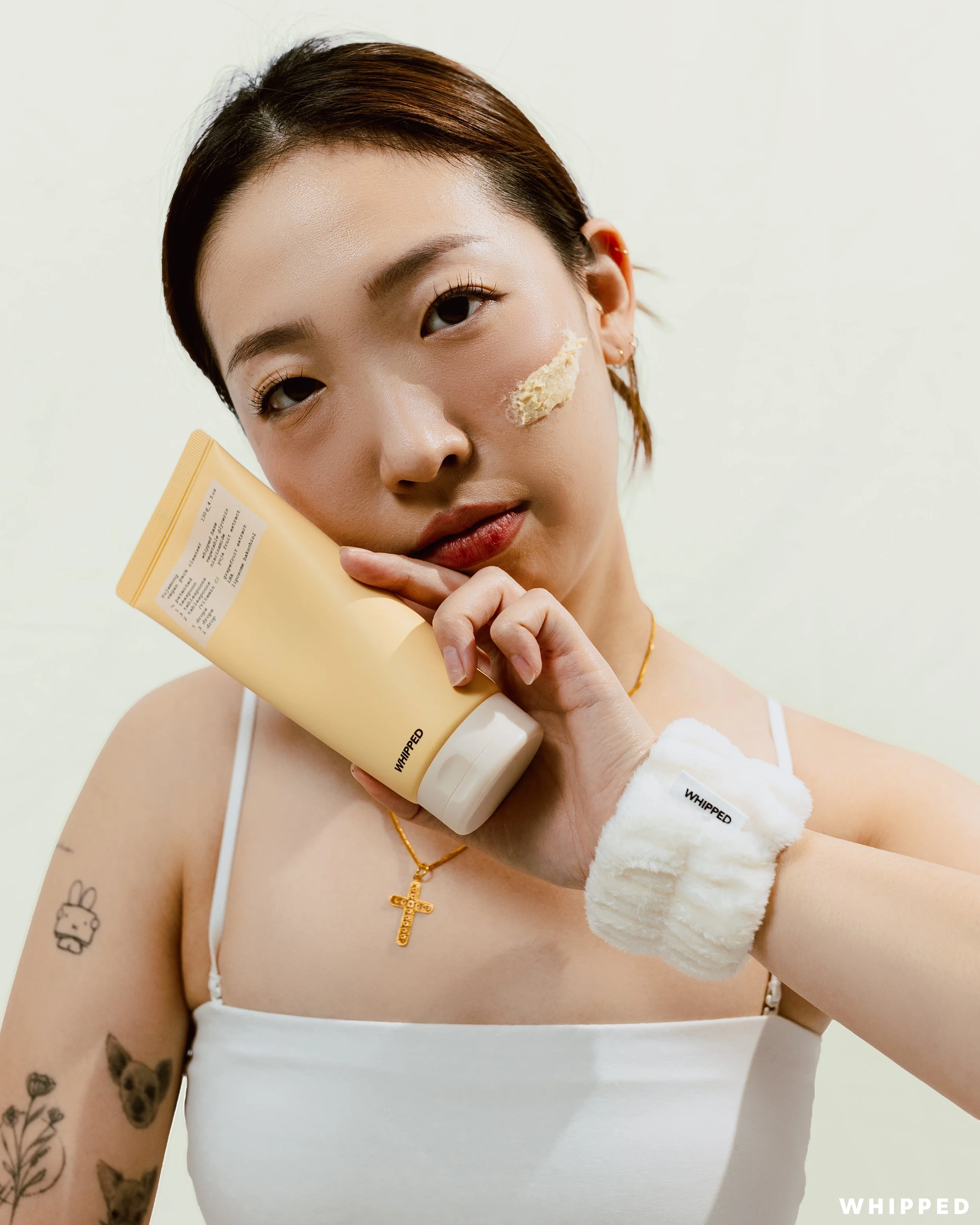Young woman with tattoos on her arm, wearing a white tank top, gold necklace with a cross, and a white fluffy wristband, holding a yellow tube of cream. She has a cream patch on her right cheek and is posing with a neutral expression against a plain 