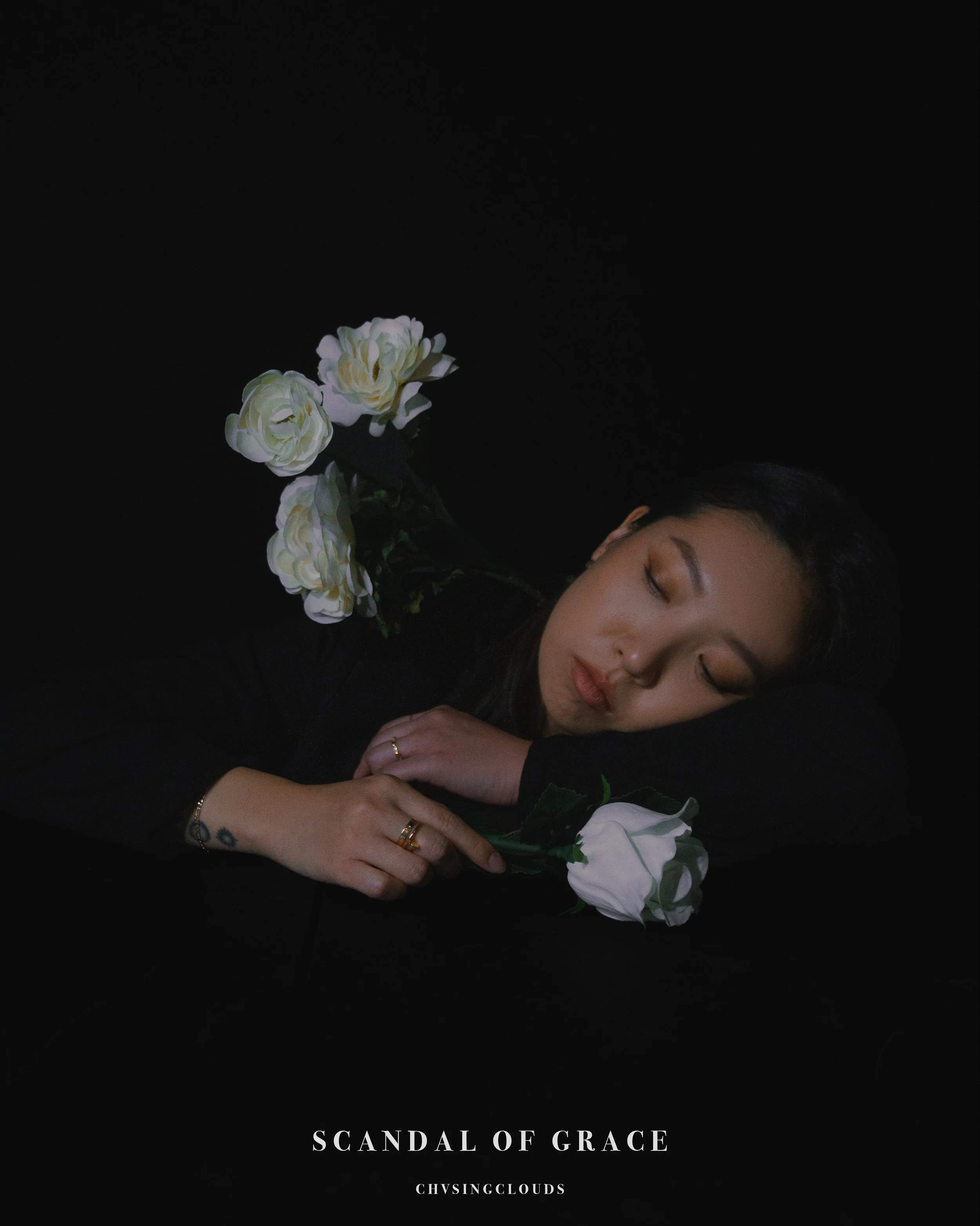 A woman with long dark hair sleeping on a black surface with white roses around her. The text 'Scandal of Grace' and 'Chvsingclouds' is at the bottom.