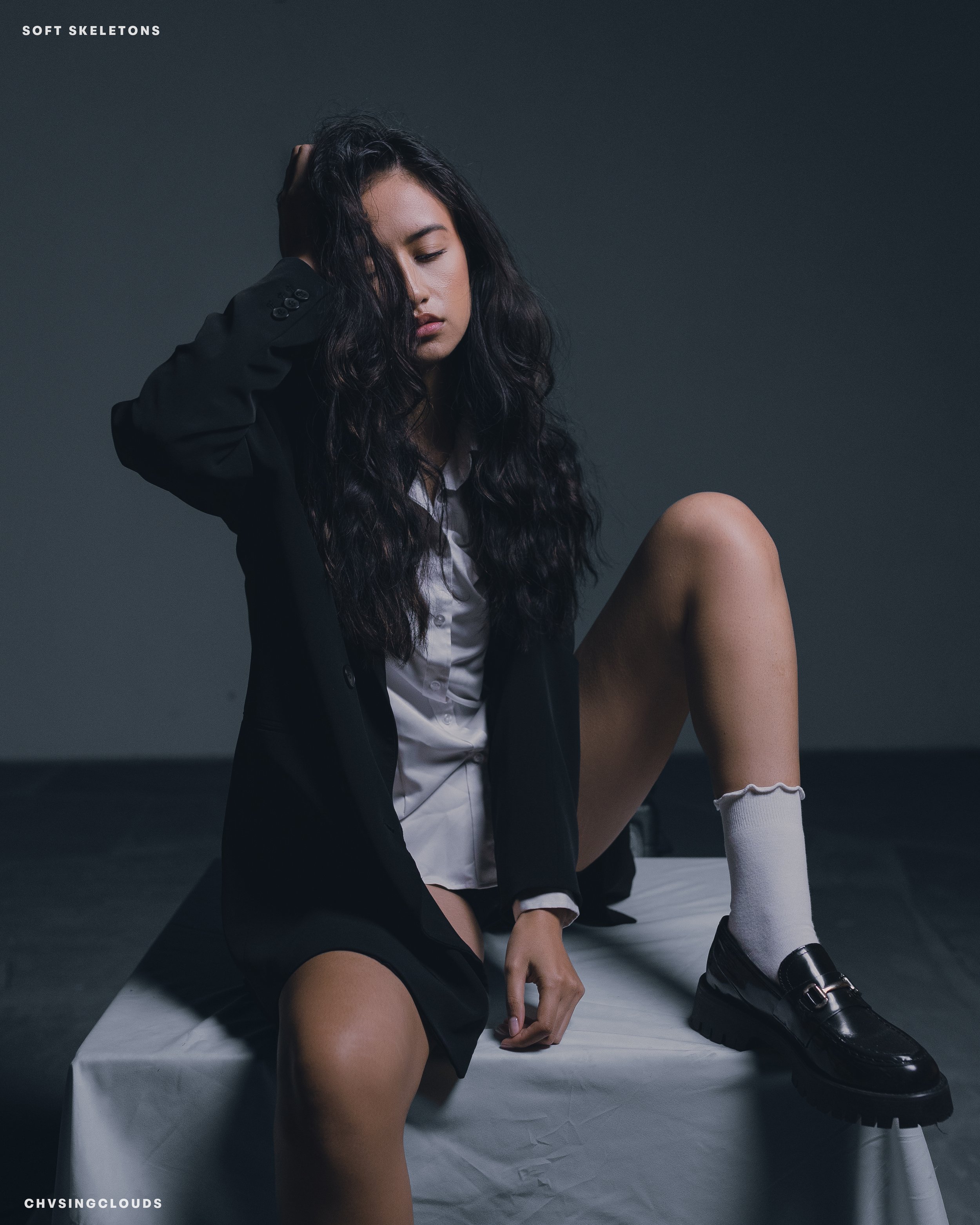 A young woman with long, wavy dark hair sitting on a white surface against a dark background. She is wearing a black blazer, white shirt, and black shoes with white socks. She has one knee raised, and her hand is touching her hair with her eyes close