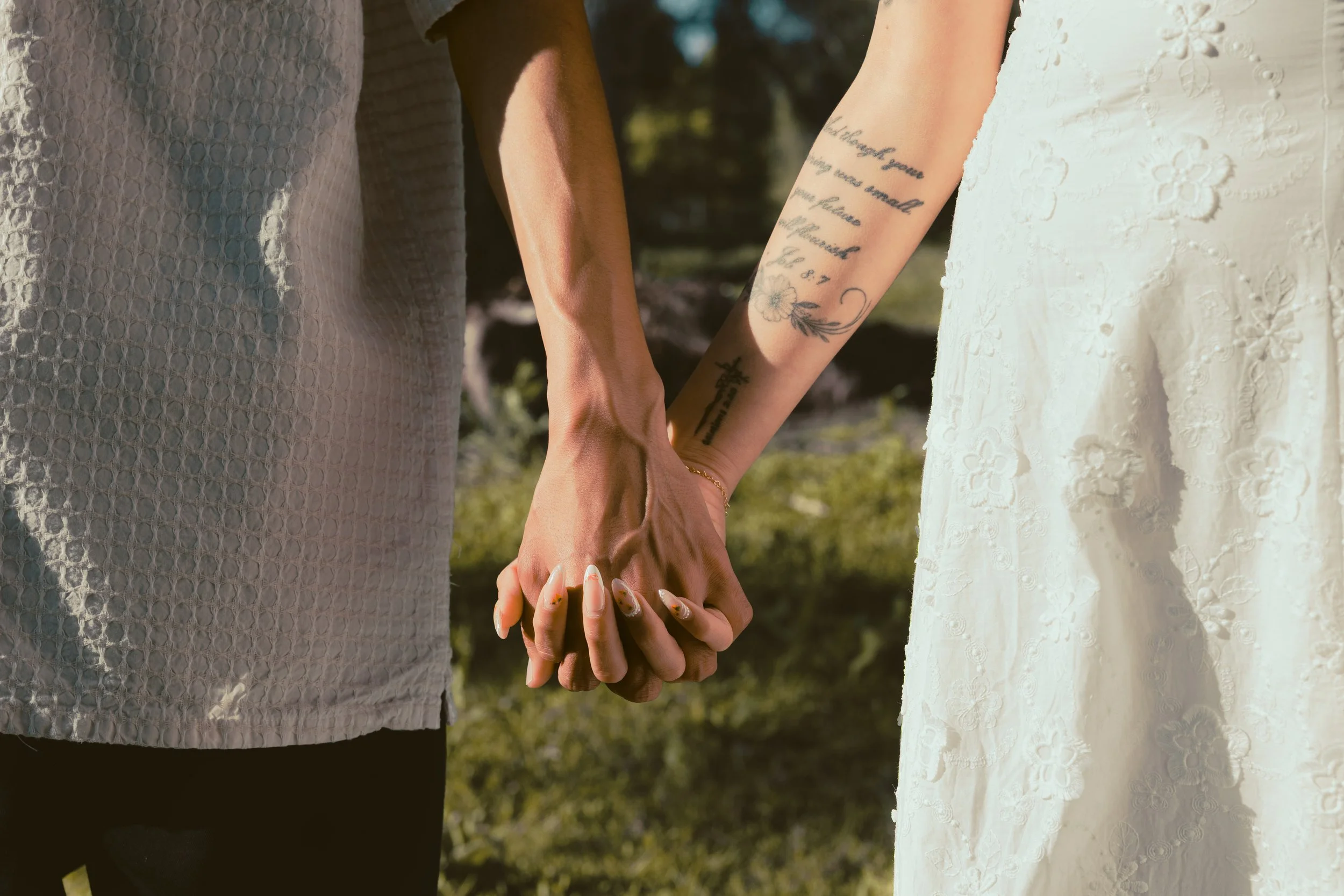 Two women holding hands, one in a grey patterned dress and the other in a white lace dress, with tattoos on their arms. They are outdoors in a grassy area.