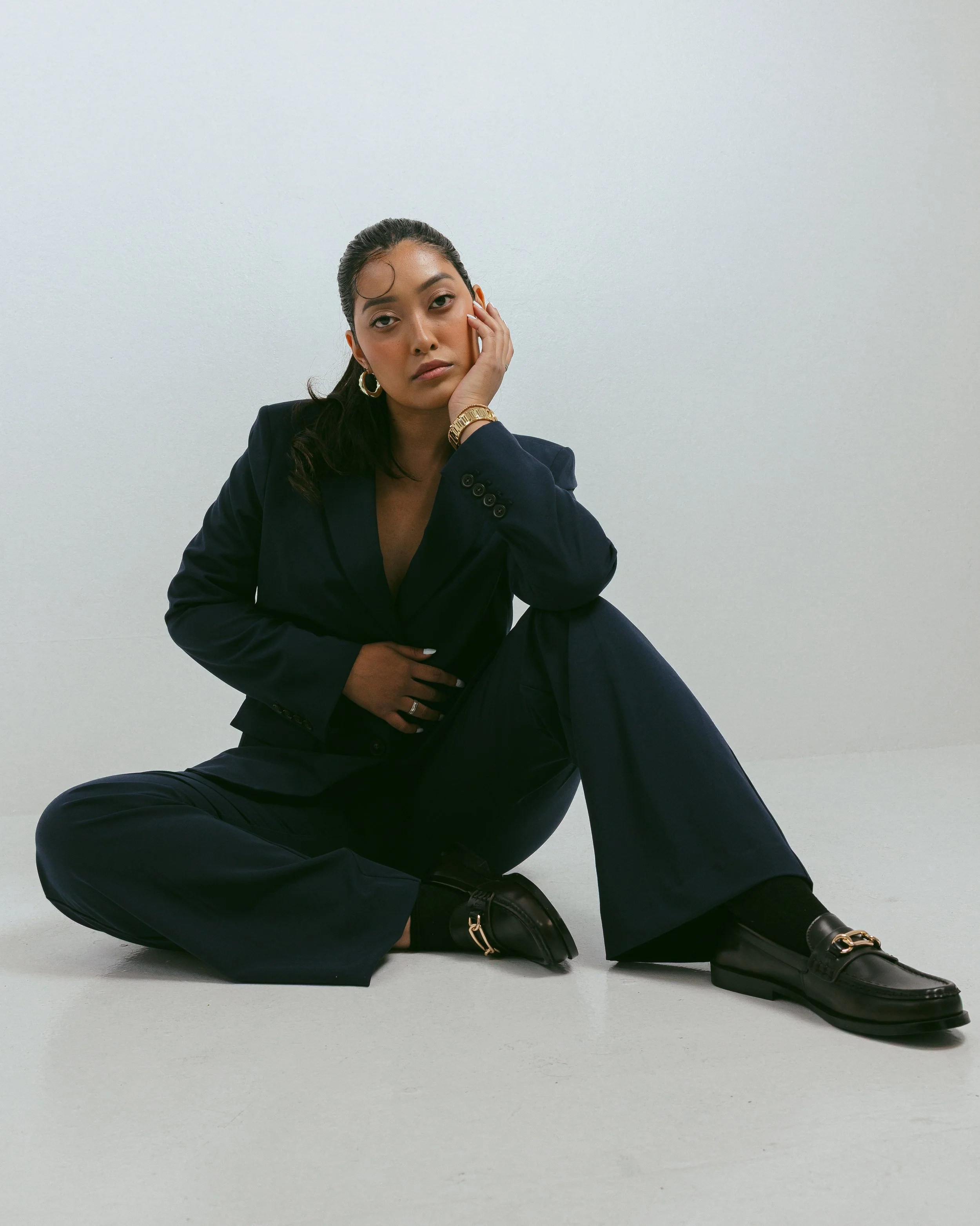 A woman in a dark suit sitting on the floor with her legs crossed in front of a plain white wall, resting her head on her right hand, with a serious expression, wearing gold jewelry and black shoes.