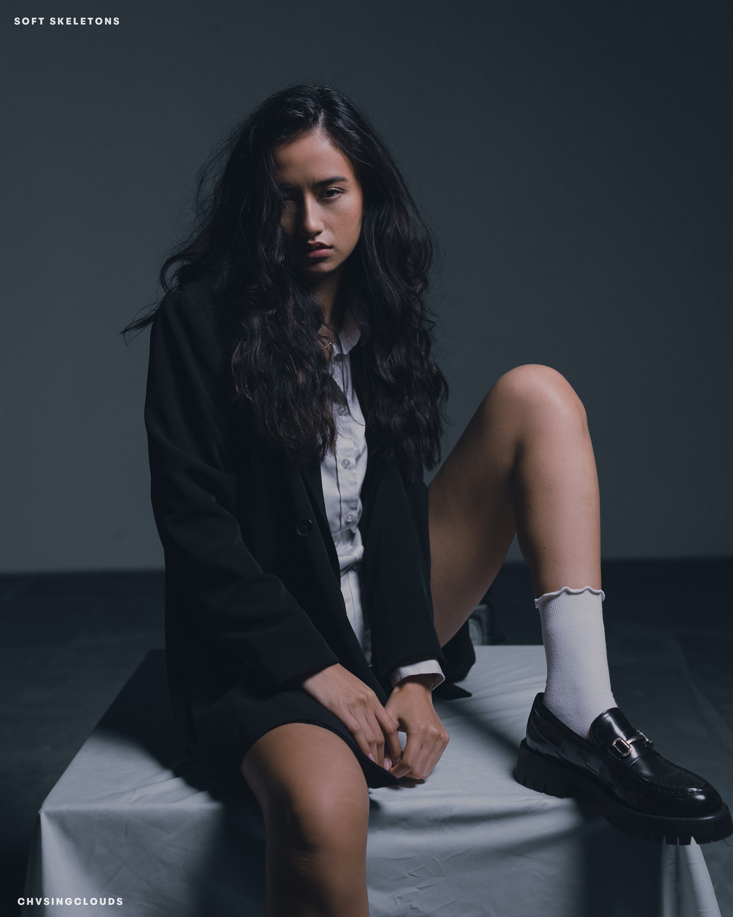 A young woman with long, wavy black hair sitting on a white table with her left leg bent and her right leg hanging down. She is wearing a black blazer, a white shirt, a black skirt, white socks, and black loafers, with a serious expression and dark b