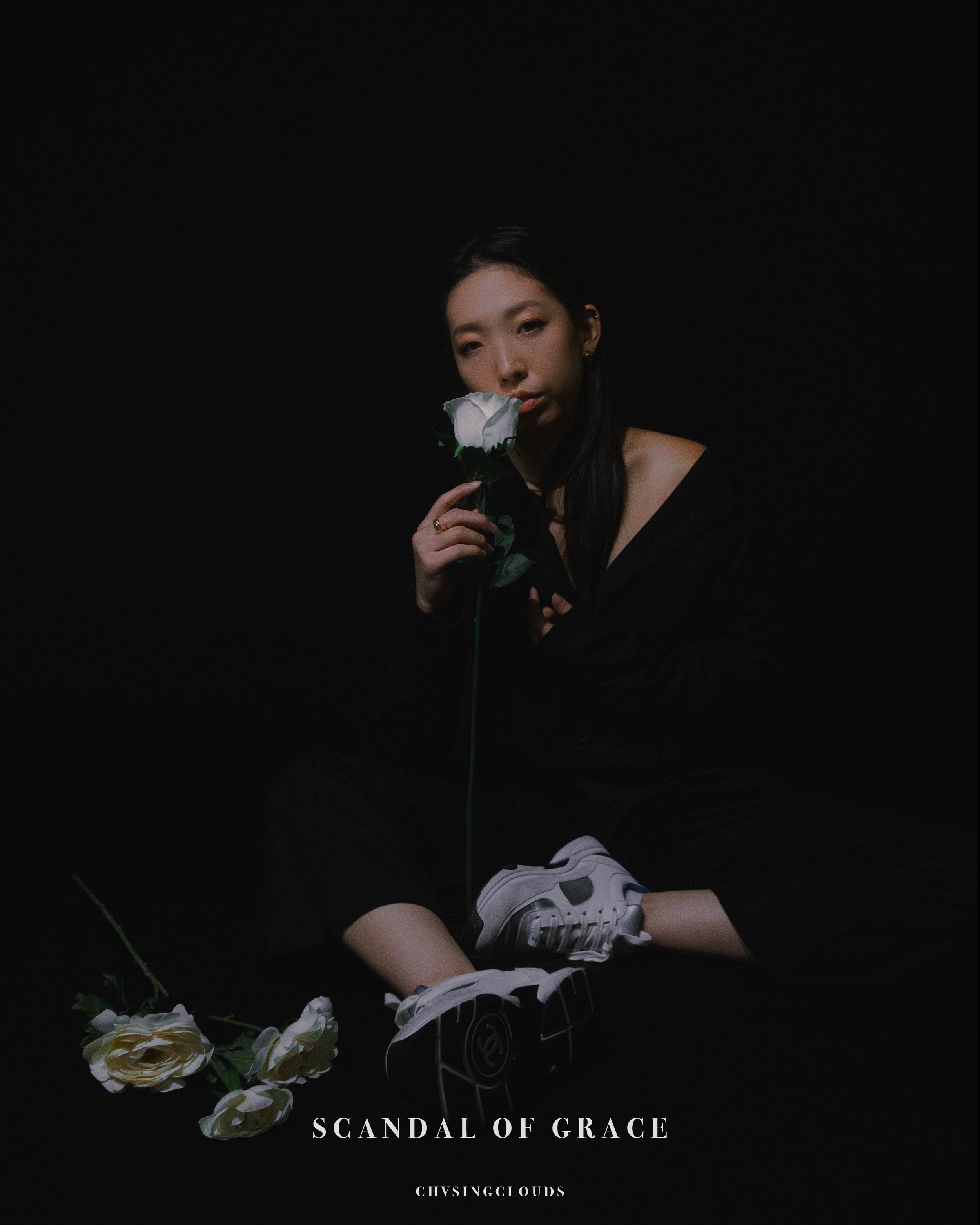 A woman in black clothing holds a white rose close to her face, sitting against a dark background, with a few white roses on the floor, and text that reads 'Scandal of Grace'.