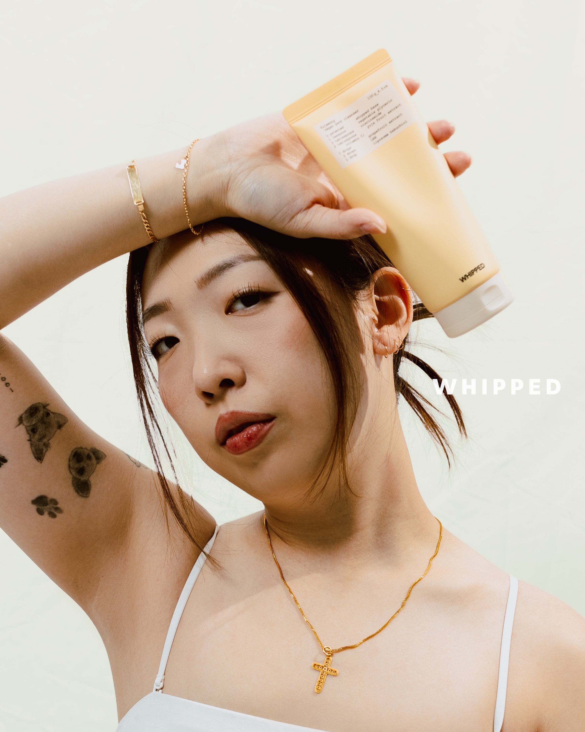 Young woman with tattoos and jewelry holding a yellow bottle on her head, looking at the camera against a plain background with the word 'WHIPPED' on the right.