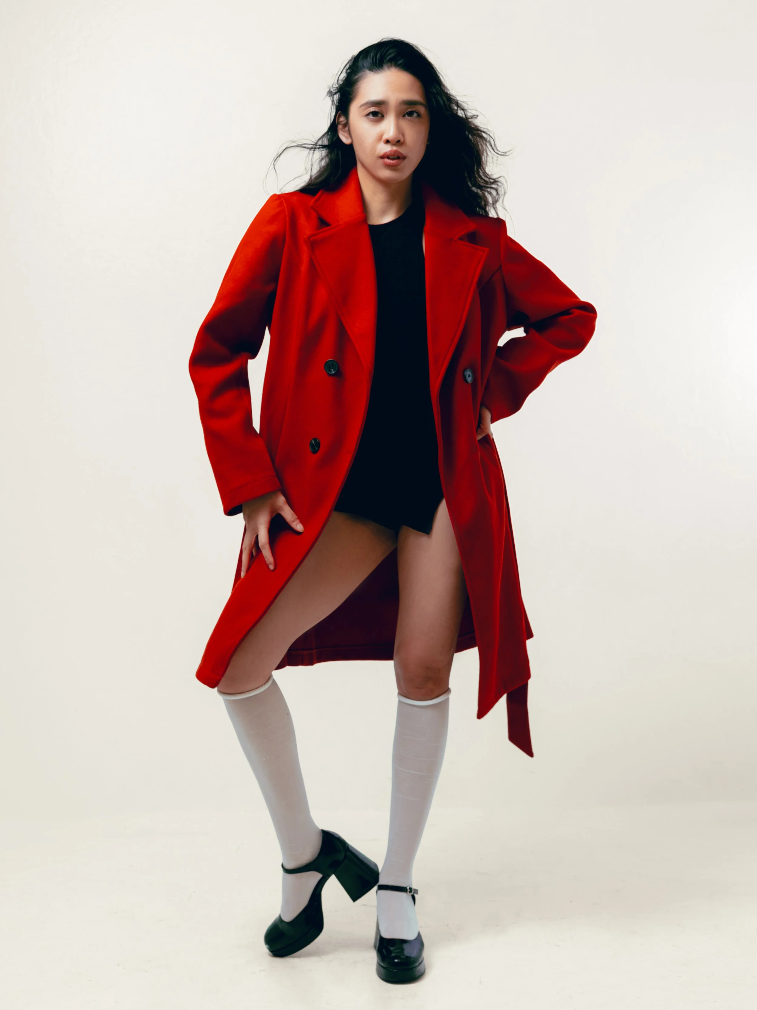 A woman wearing a red coat, black top, white thigh-high socks, and black shoes, posing with one hand on her hip and the other holding her coat open against a plain white background.