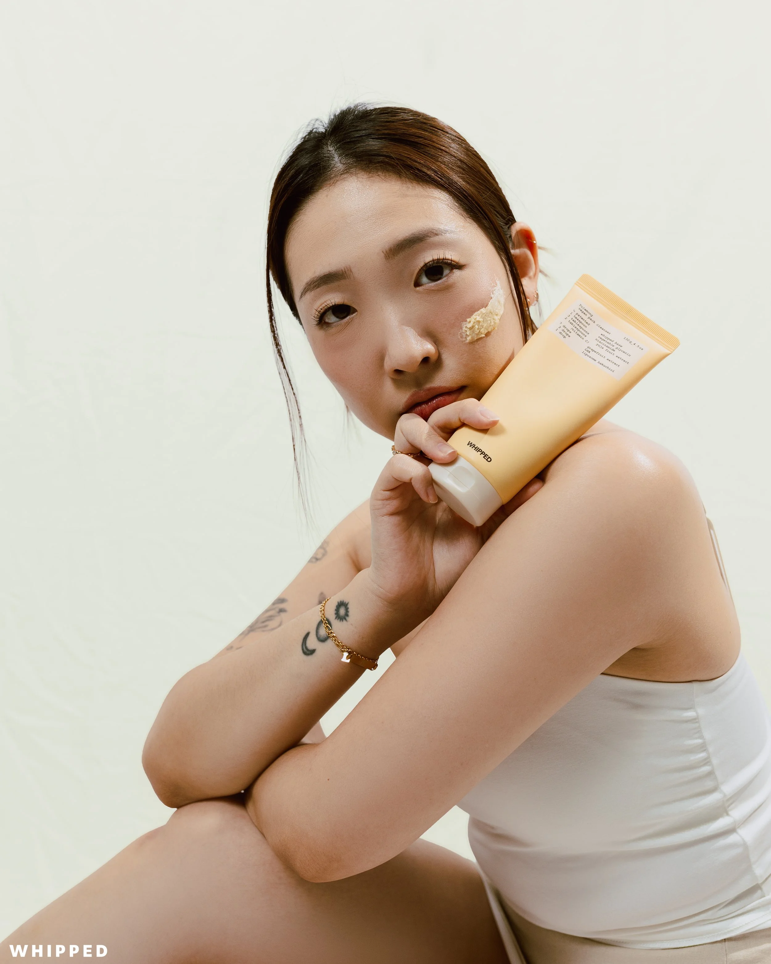 A young woman with tattoos on her arm is holding a tube of skincare product close to her face. She is wearing a white sleeveless top and has a patch of the product on her cheek, with a neutral expression, against a plain light background.
