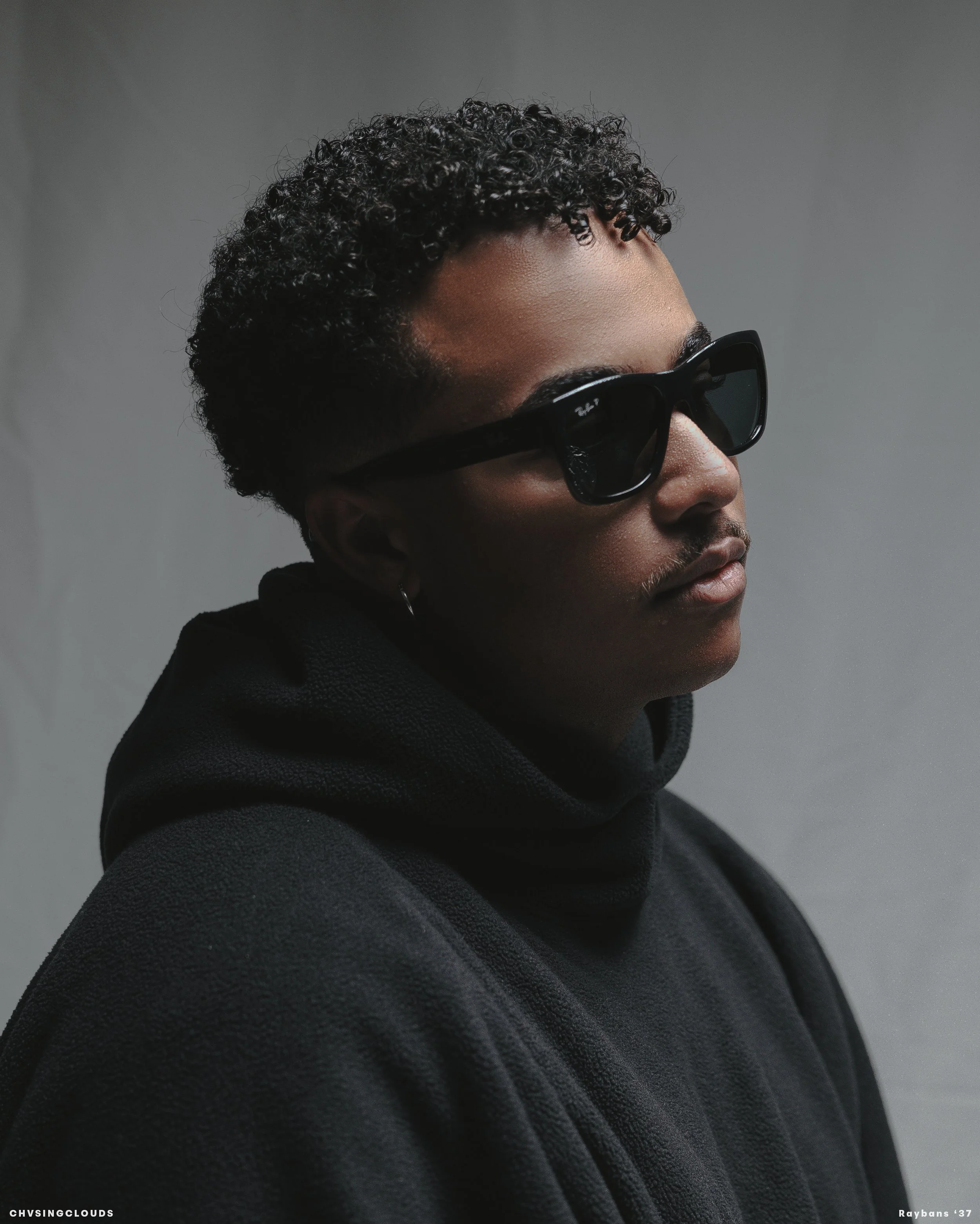 A person with curly black hair wearing dark sunglasses and a black hoodie, posing against a gray background.