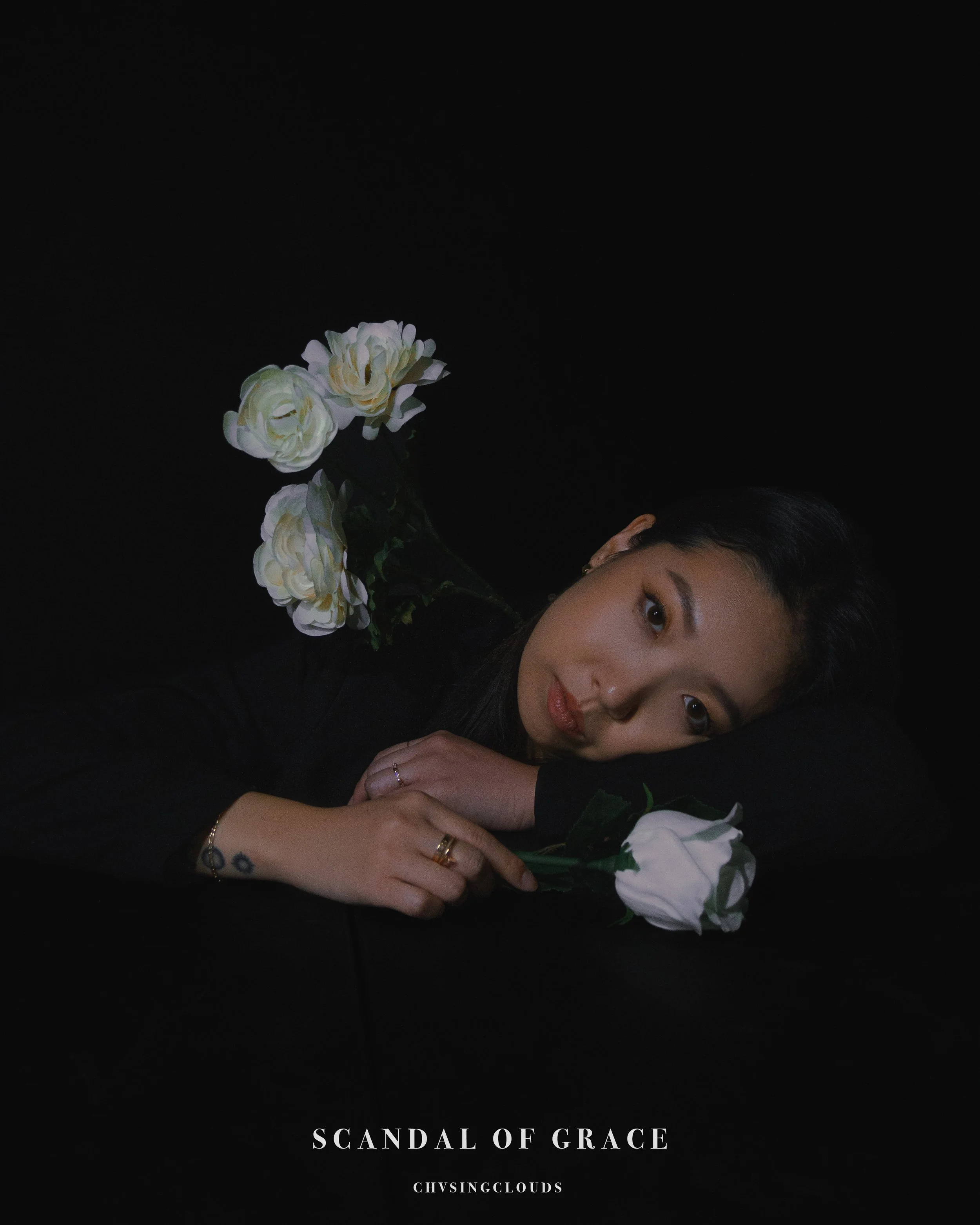 A woman with dark hair lying on her side on a black surface, resting her head on her arm, with three white roses around her. The background is black, and there is text that reads 'Scandal of Grace' and 'Chvsingclouds'.