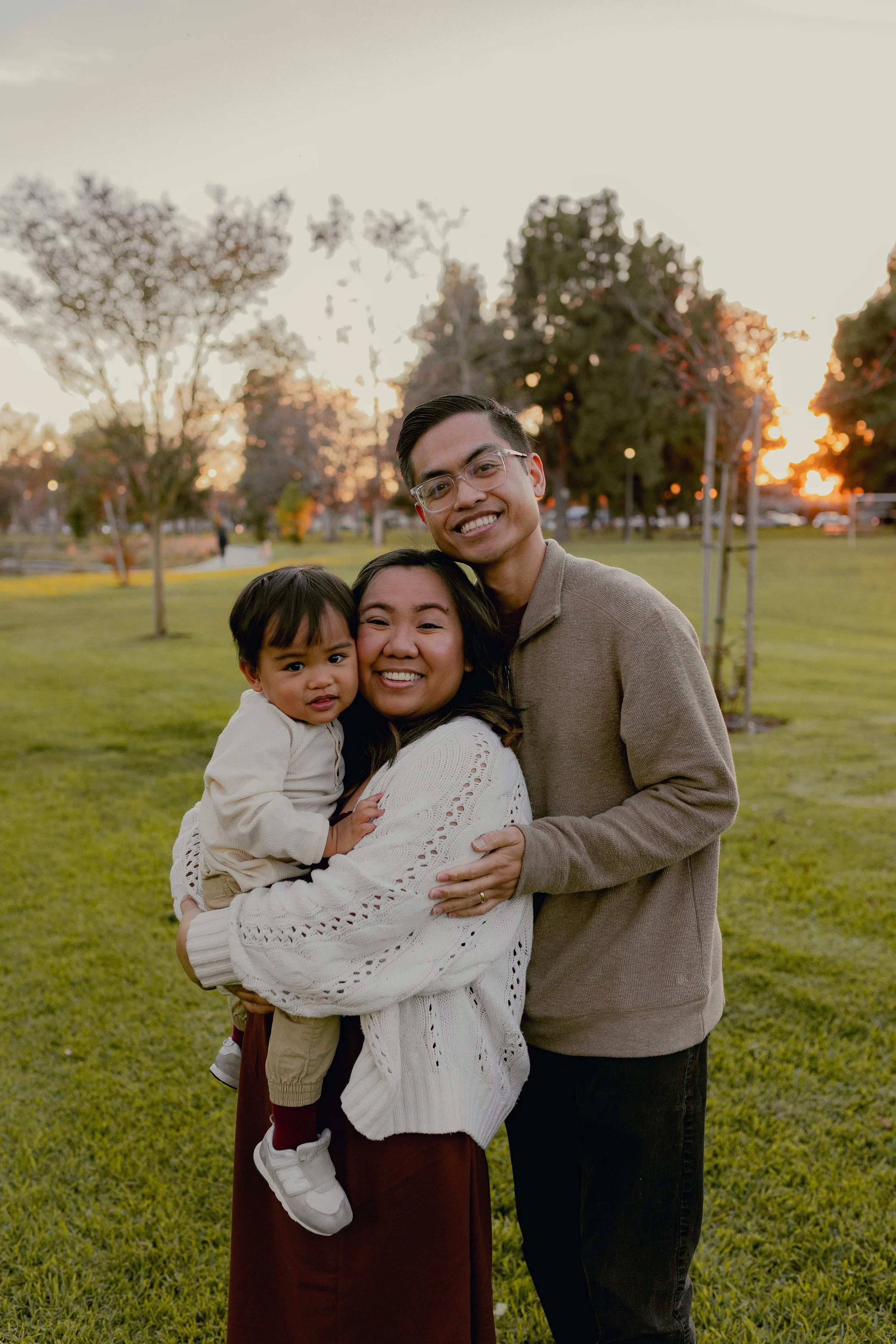 A family of three smiling and hugging outdoors during sunset, with trees and a park in the background.