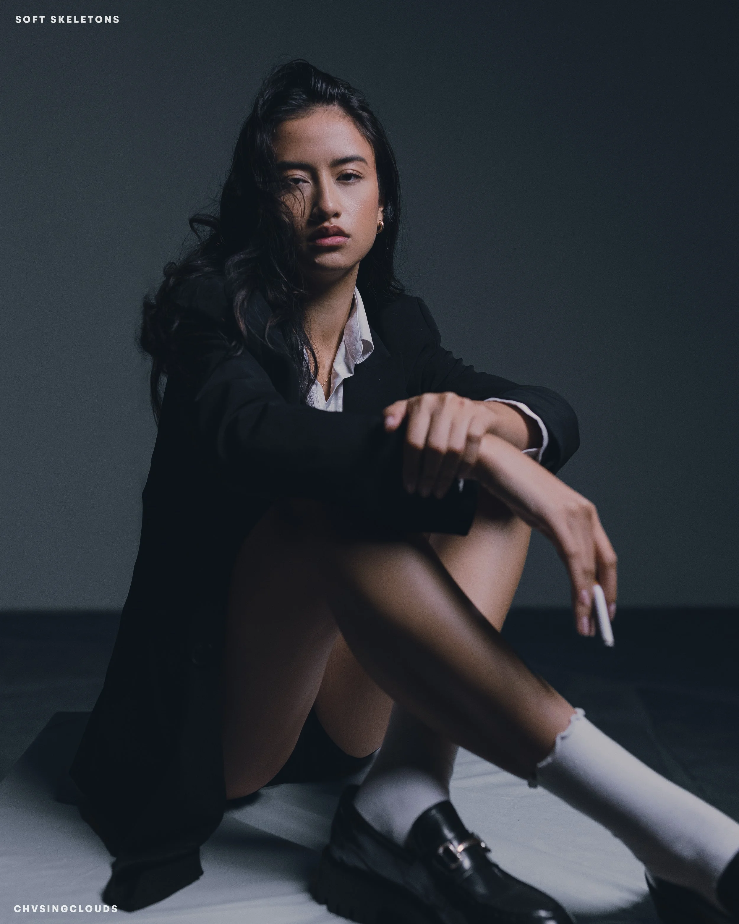 A young woman with dark wavy hair sitting on the floor against a plain dark background, wearing a black blazer, white shirt, black shoes, and white socks, with her left arm resting on her knee.