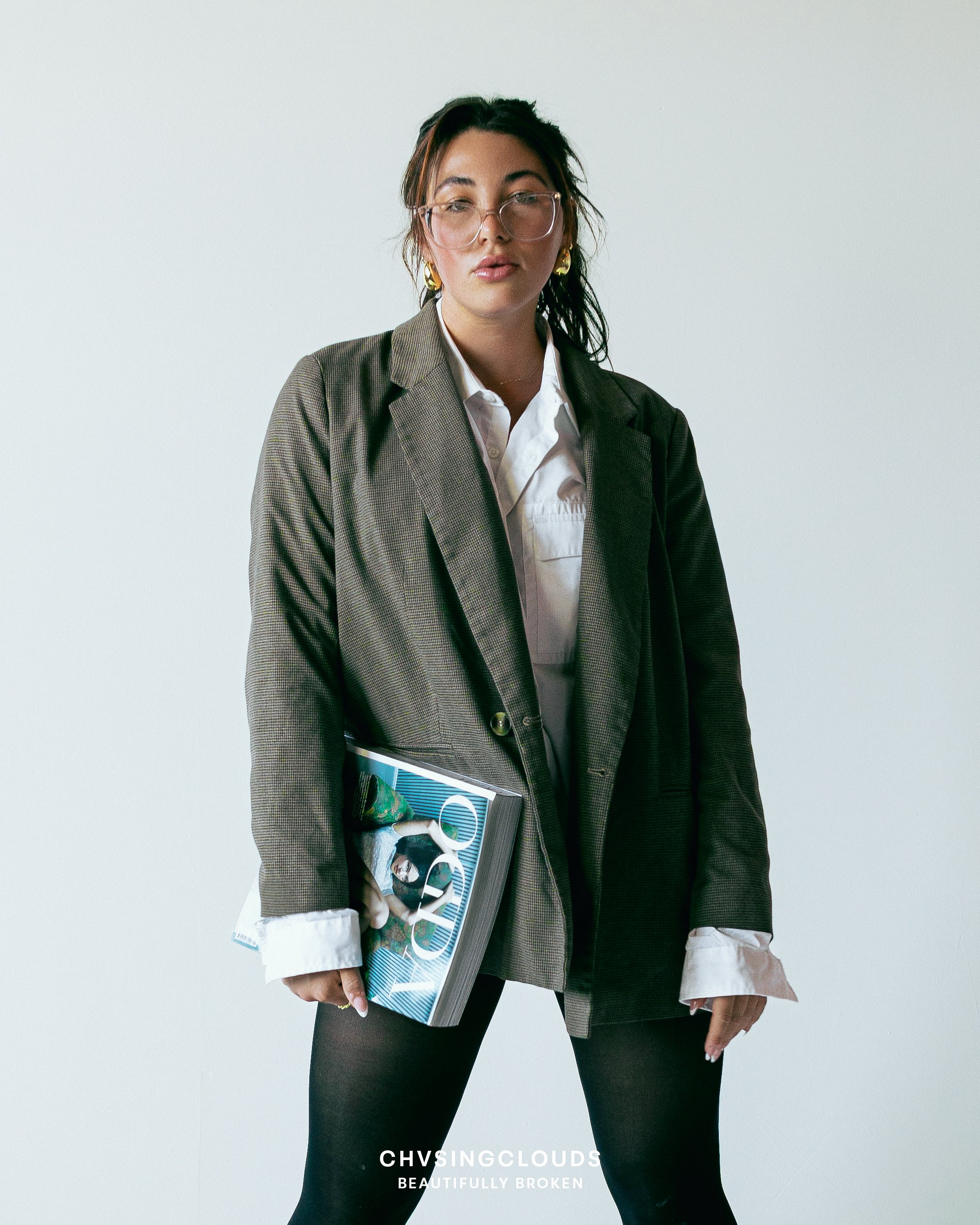 A woman with glasses, earrings, and dark hair wearing a brown blazer over a white shirt, holding a magazine, standing against a plain light background.