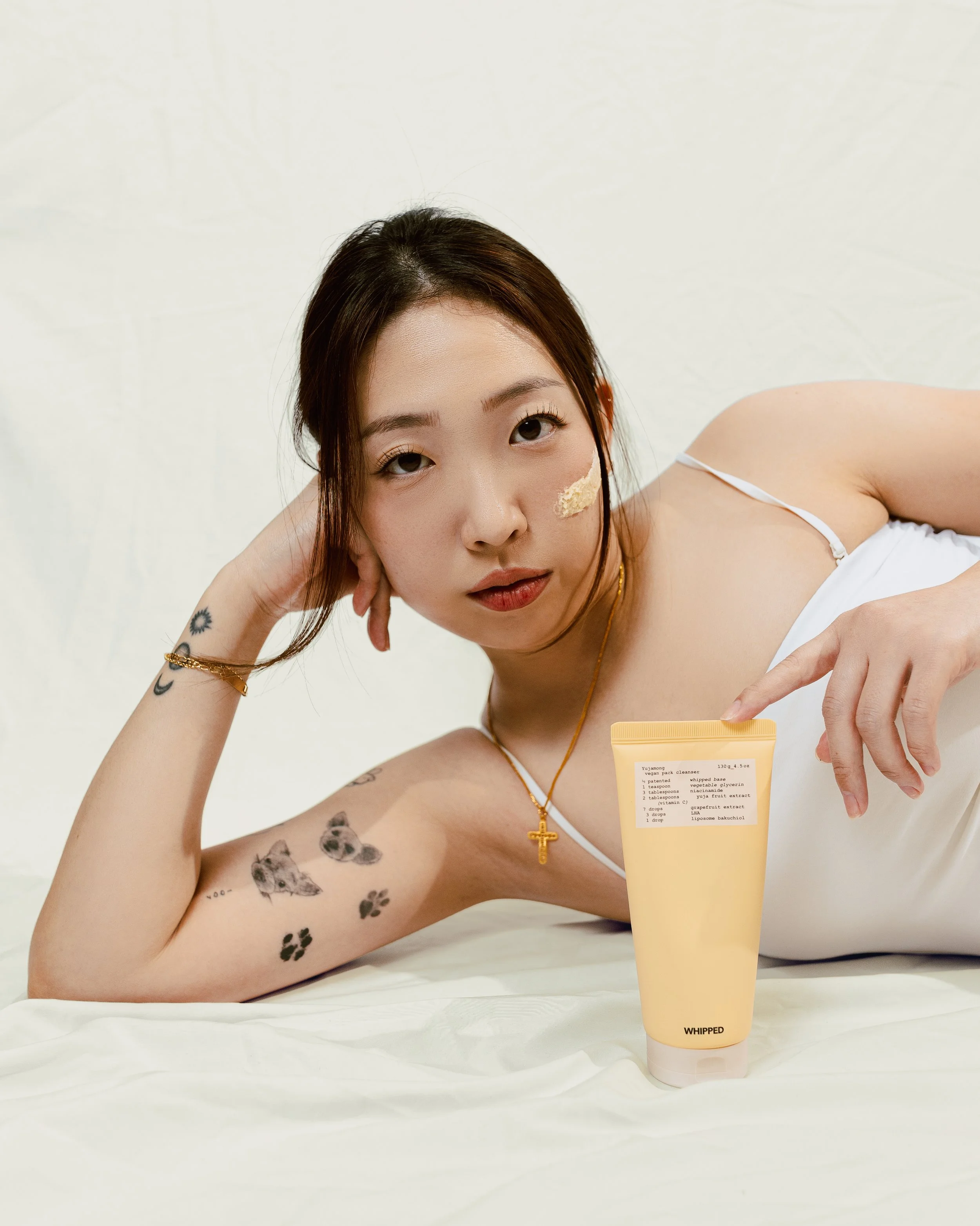 A young woman with tattoos lying on her side on a bed, resting her head on her hand, pointing at a yellow skincare product in front of her, with a neutral expression.