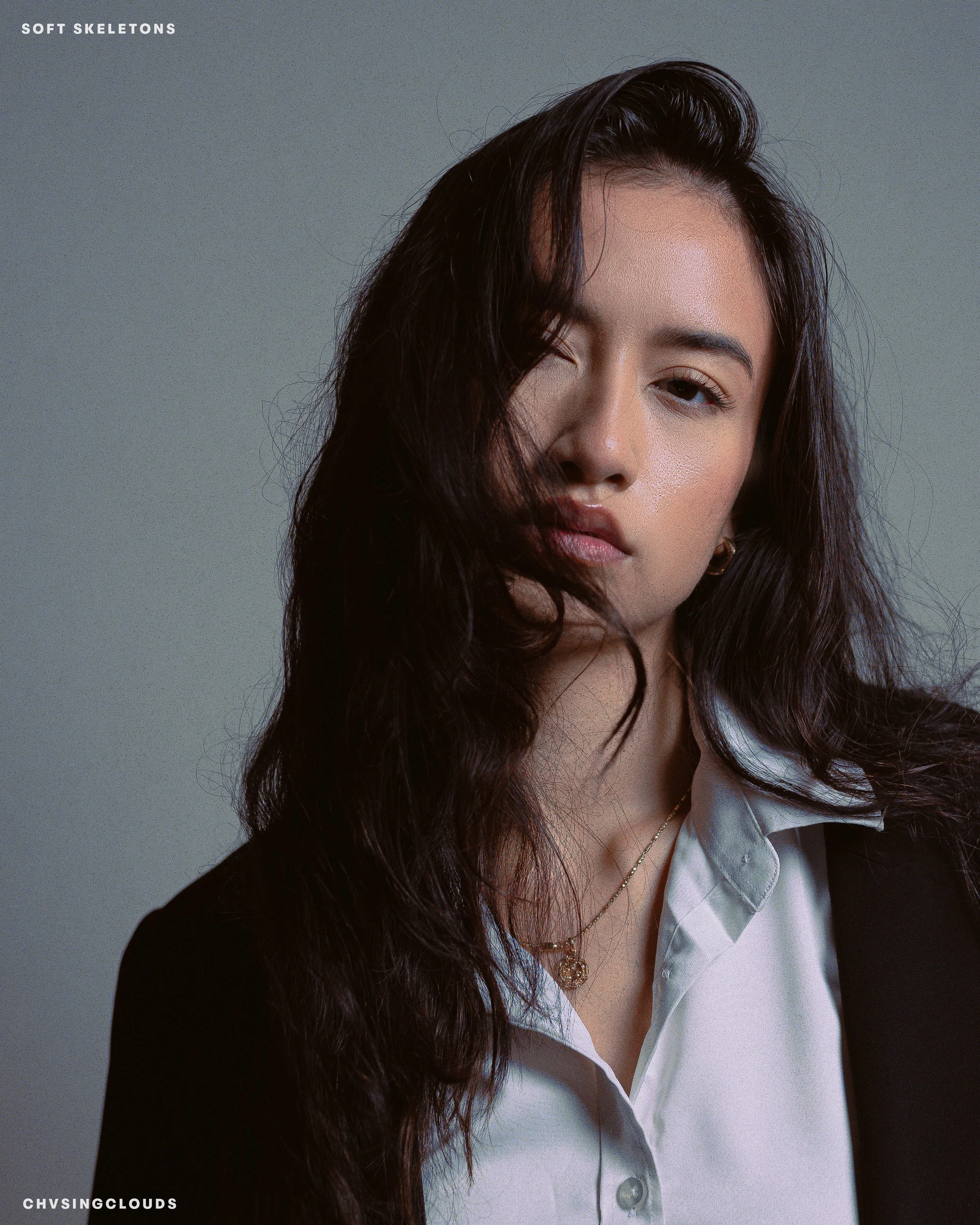 Close-up of a woman with dark, wavy hair wearing a white collared shirt and a black blazer, looking at the camera with a relaxed expression.