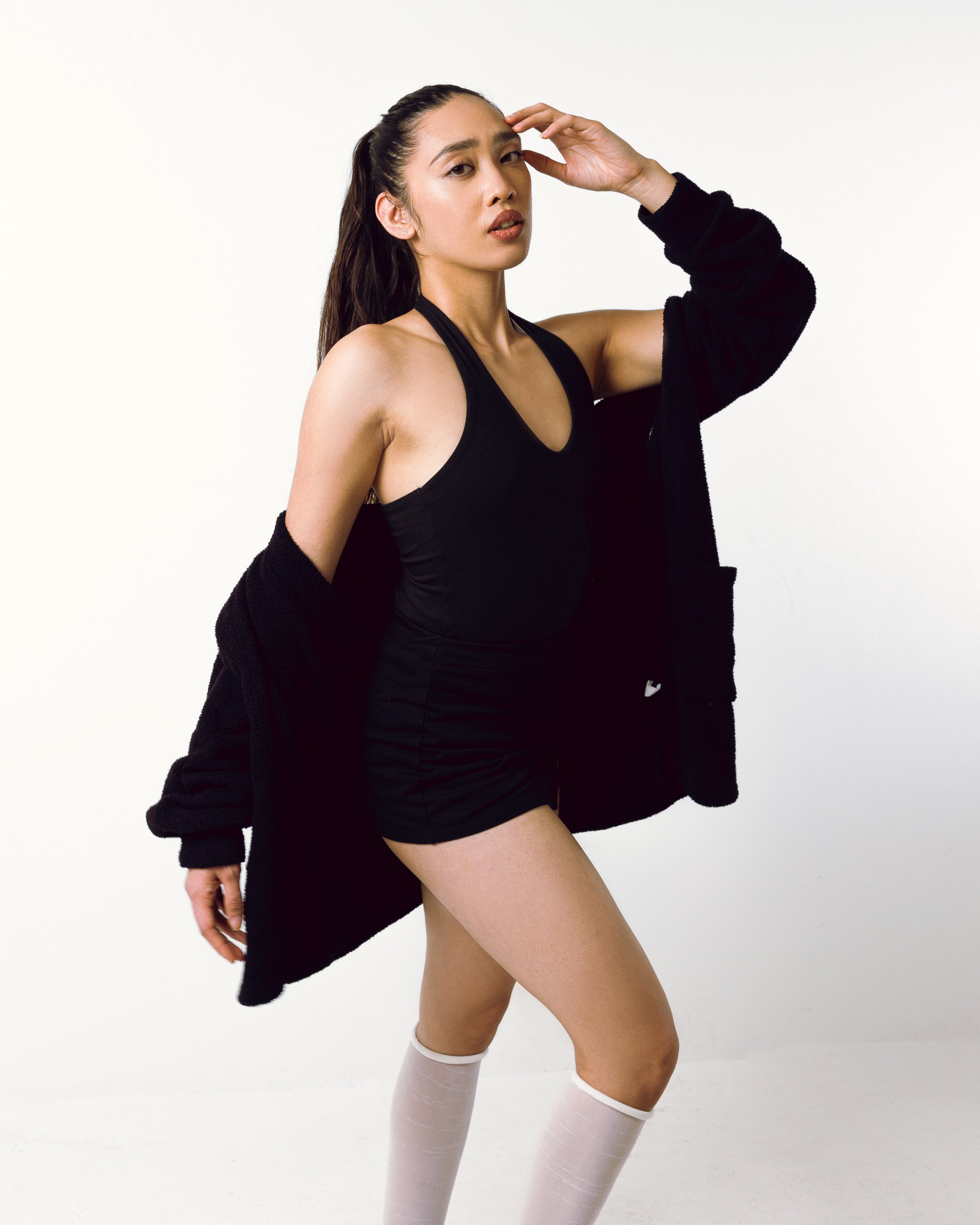 A young woman with long dark hair, wearing a black athletic tank top, black shorts, a black oversized sweater, and white knee-high socks, standing against a white background.