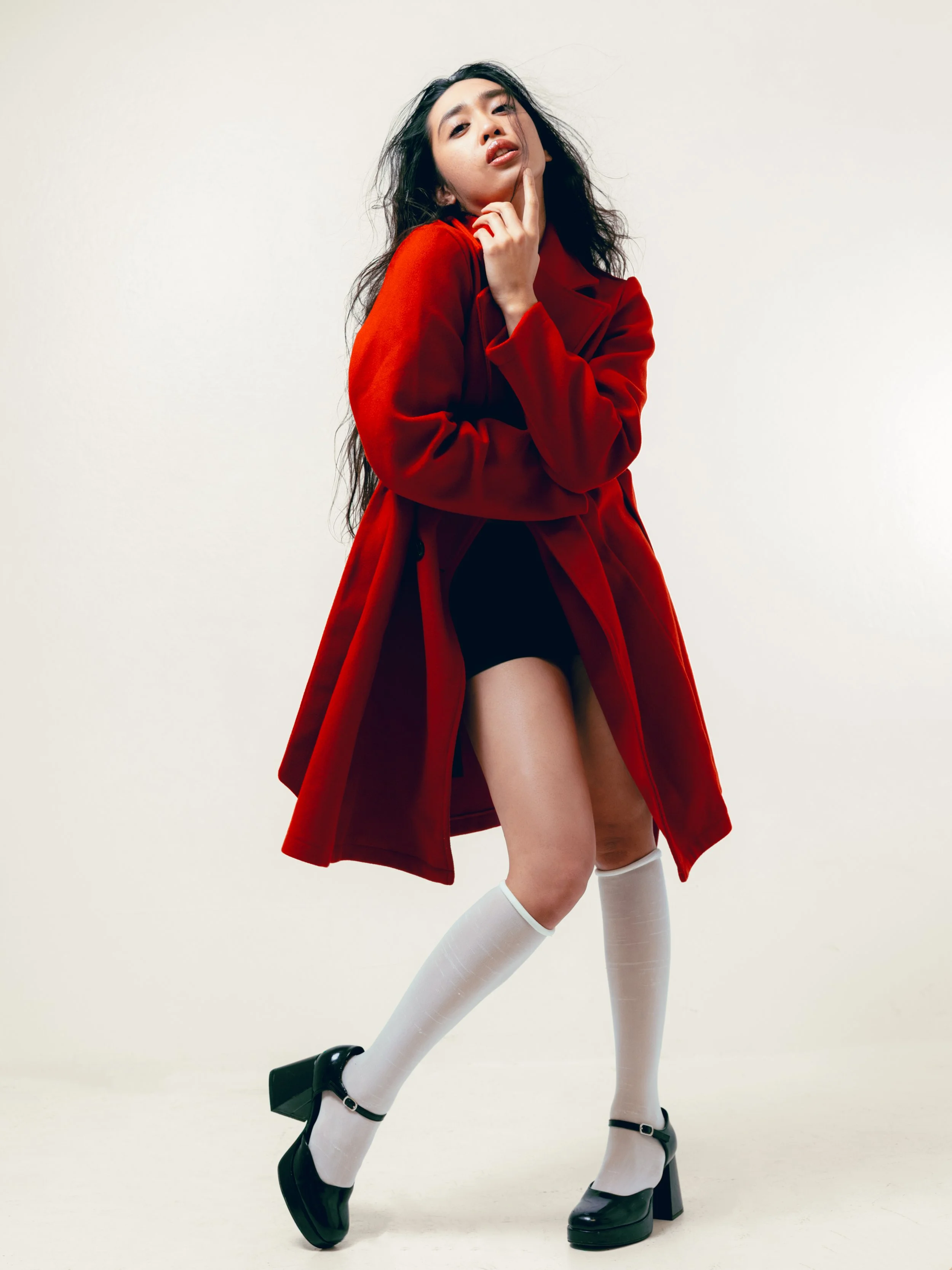 Young woman with long dark hair posing confidently, wearing a red coat, black dress, white knee-high socks, and black platform shoes, against a plain white background.