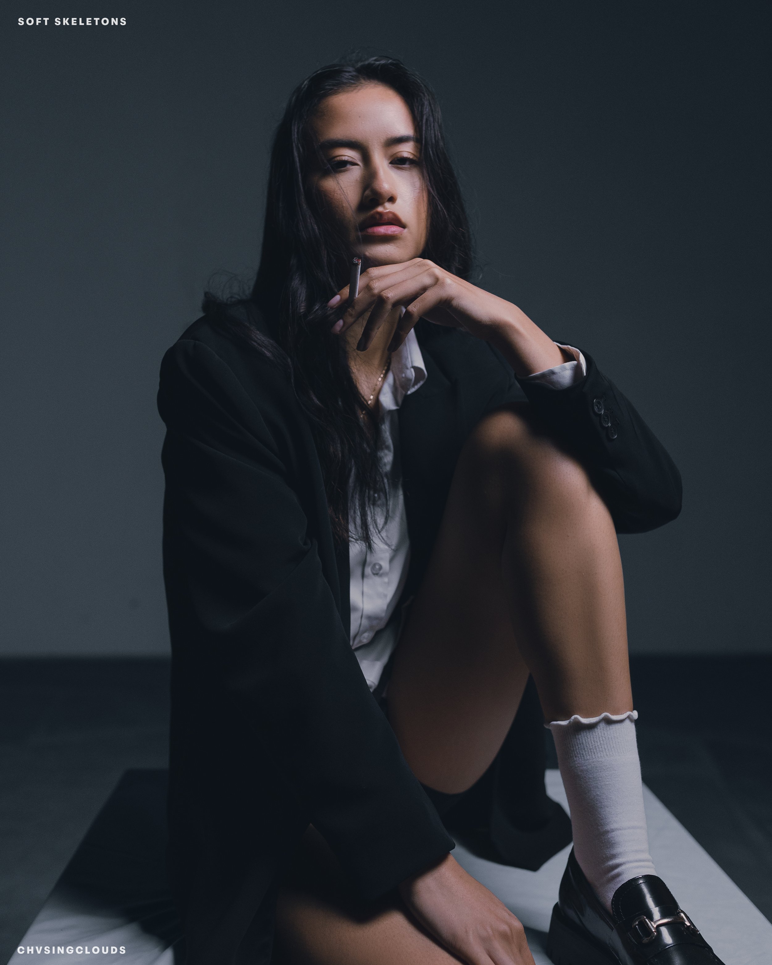 A young woman with long dark hair sitting on the floor in a dark room, wearing a black blazer, white shirt, shorts, white knee-high socks, and black shoes. She is looking at the camera with a serious expression, resting her chin on her hand, with a c