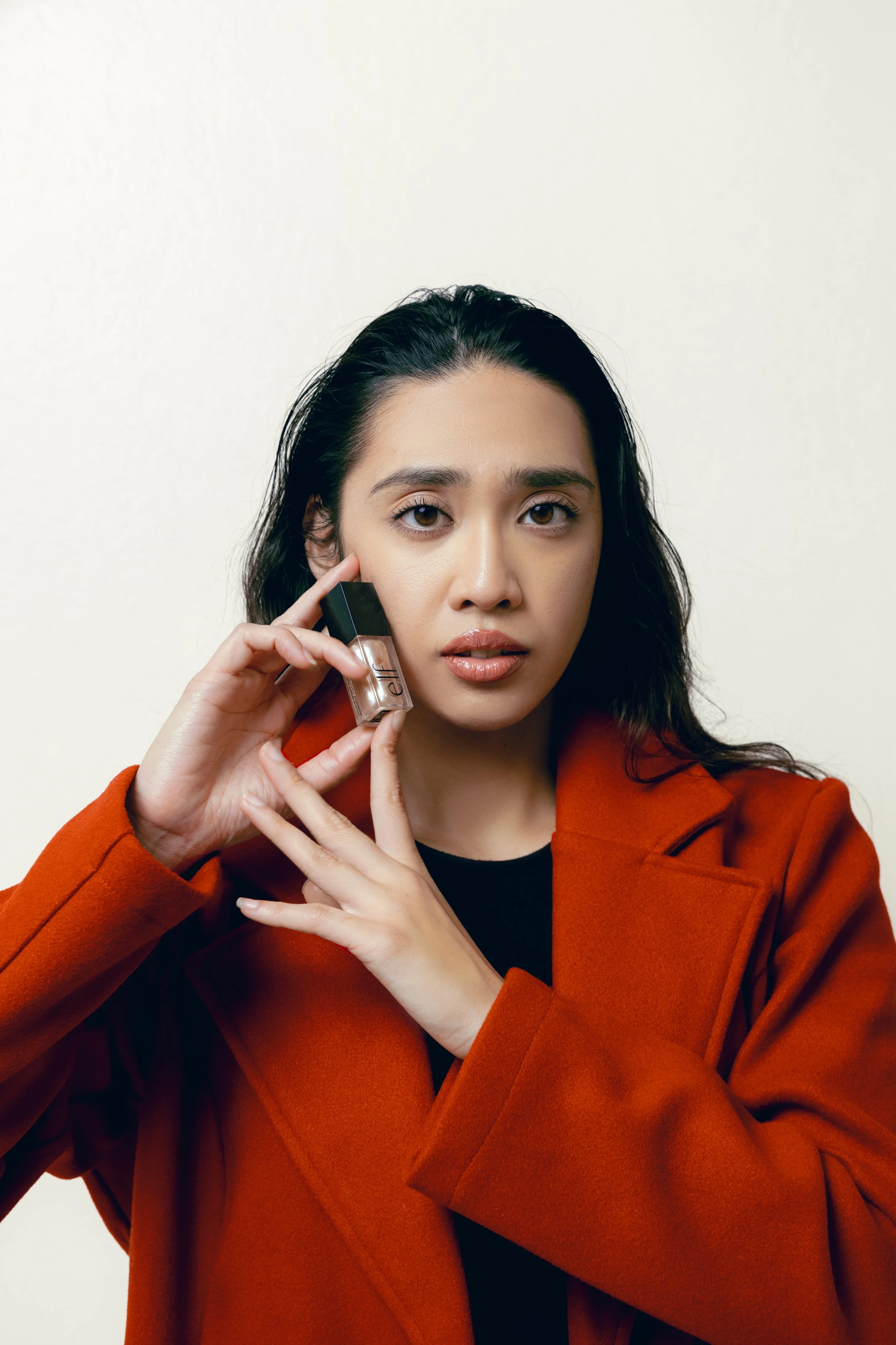 A woman with dark hair and light skin, wearing a rust-colored coat, holding a small bottle of Elle nail polish near her face, looking directly at the camera with a neutral expression, against a plain light background.