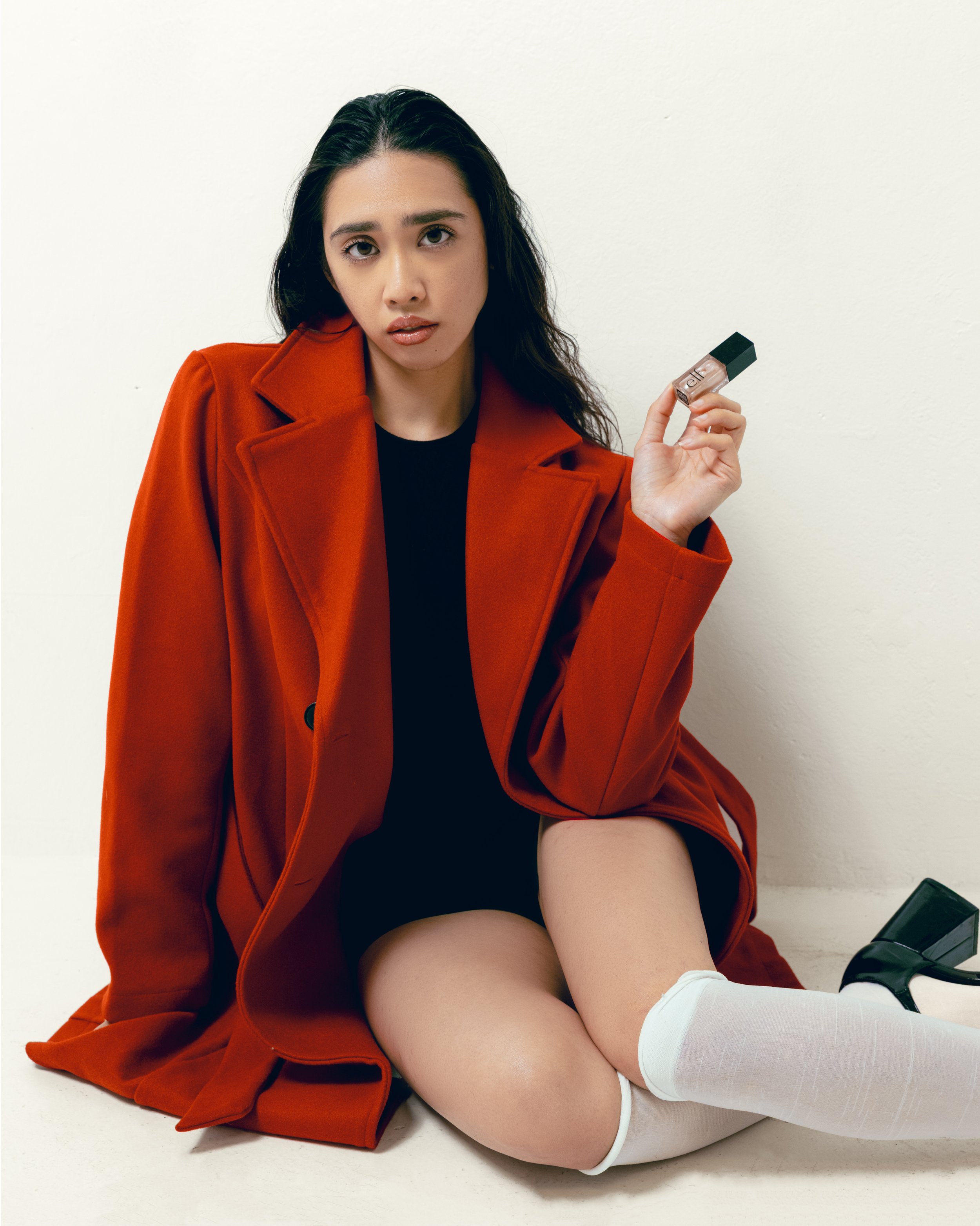 A woman sitting on the floor wearing a red coat, black top, white thigh-high socks, and black shoes, holding a small makeup product against a plain white wall.