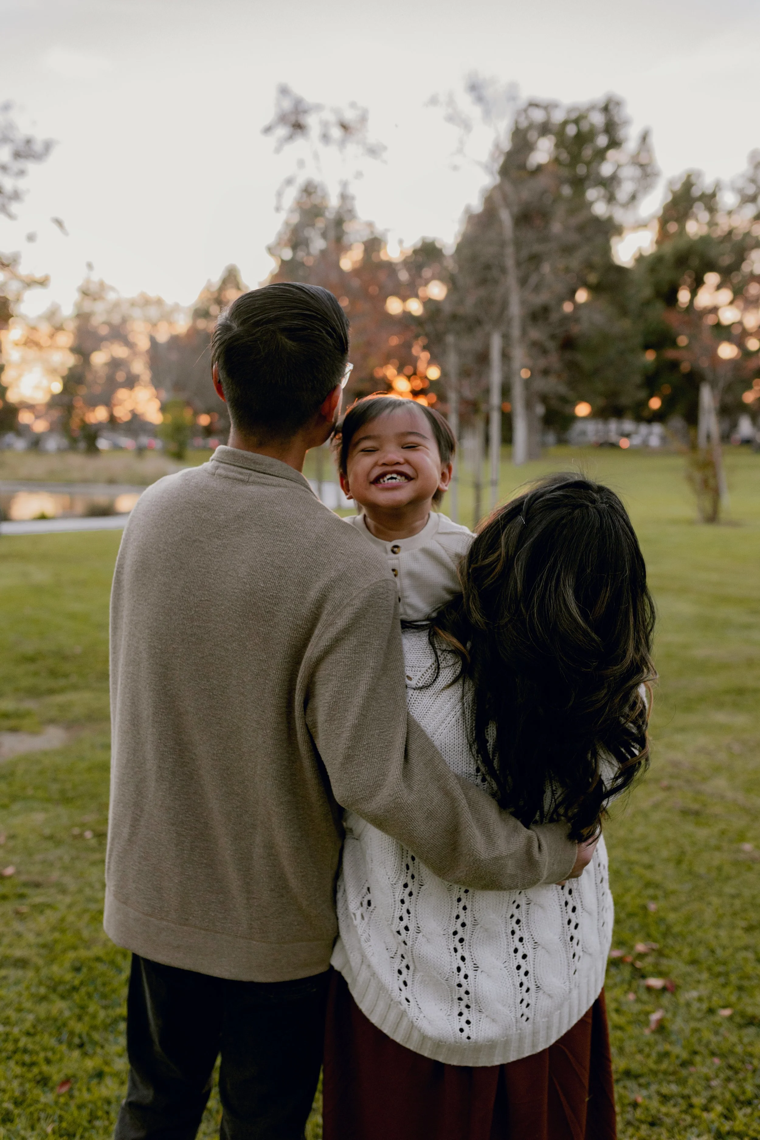 A family of three enjoying an outdoor moment during sunset, with a young girl smiling and being held by her parents in a park.