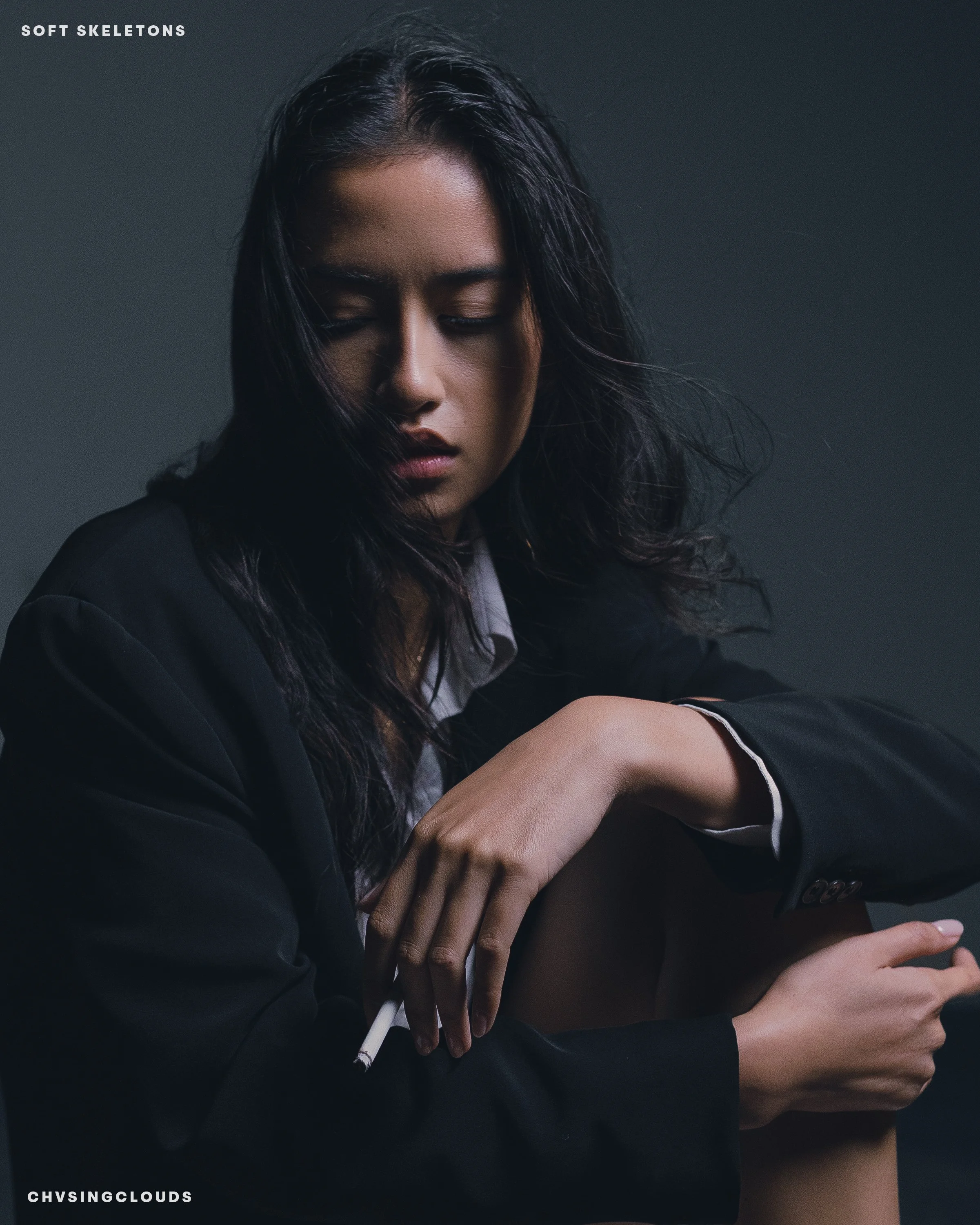 A woman with long dark hair wearing a black blazer and holding a cigarette, with her eyes closed against a dark background.