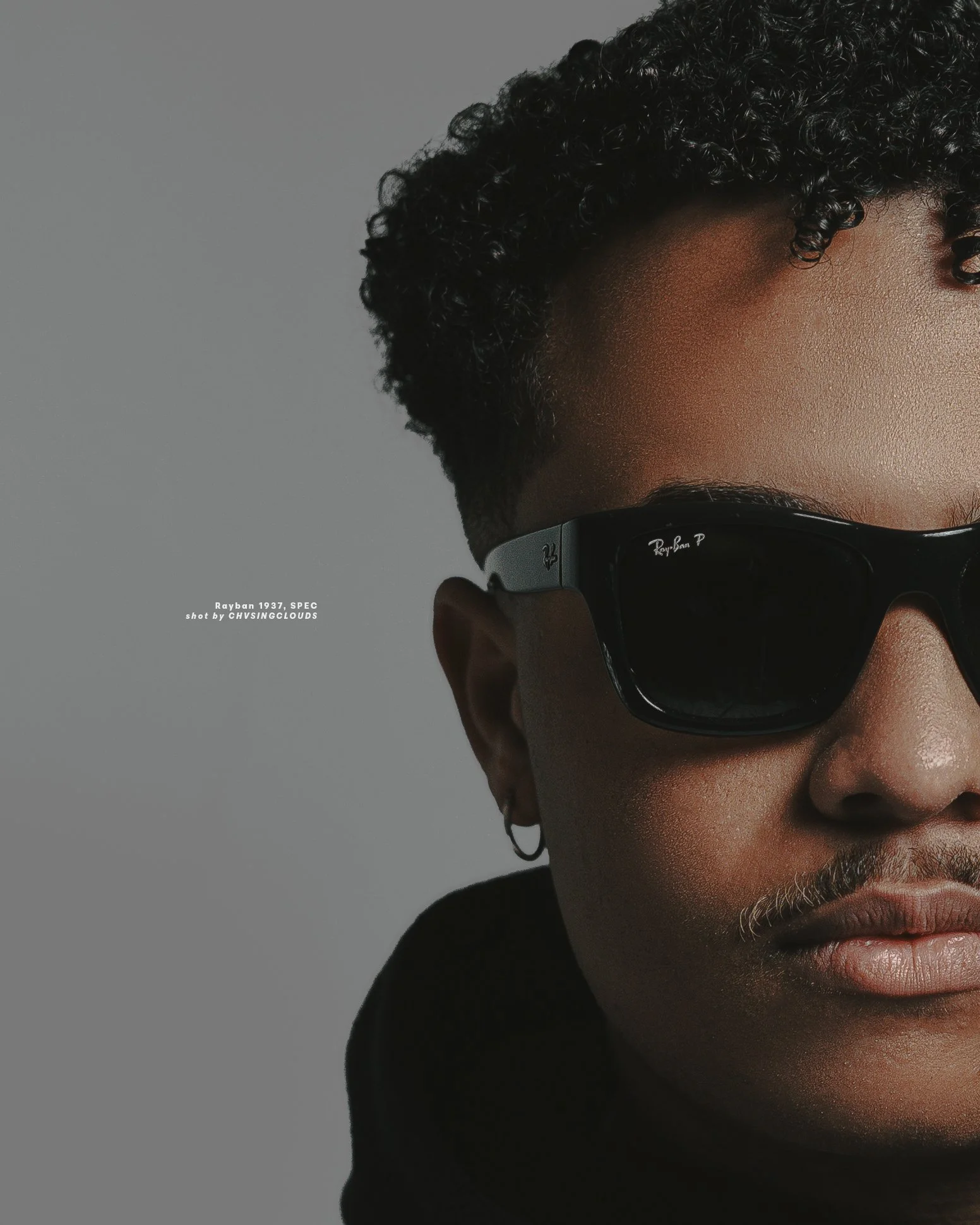 Close-up of a person wearing black Ray-Ban sunglasses, showing only part of their face with detailed skin texture, curly hair, and small hoop earrings. The background is plain gray.