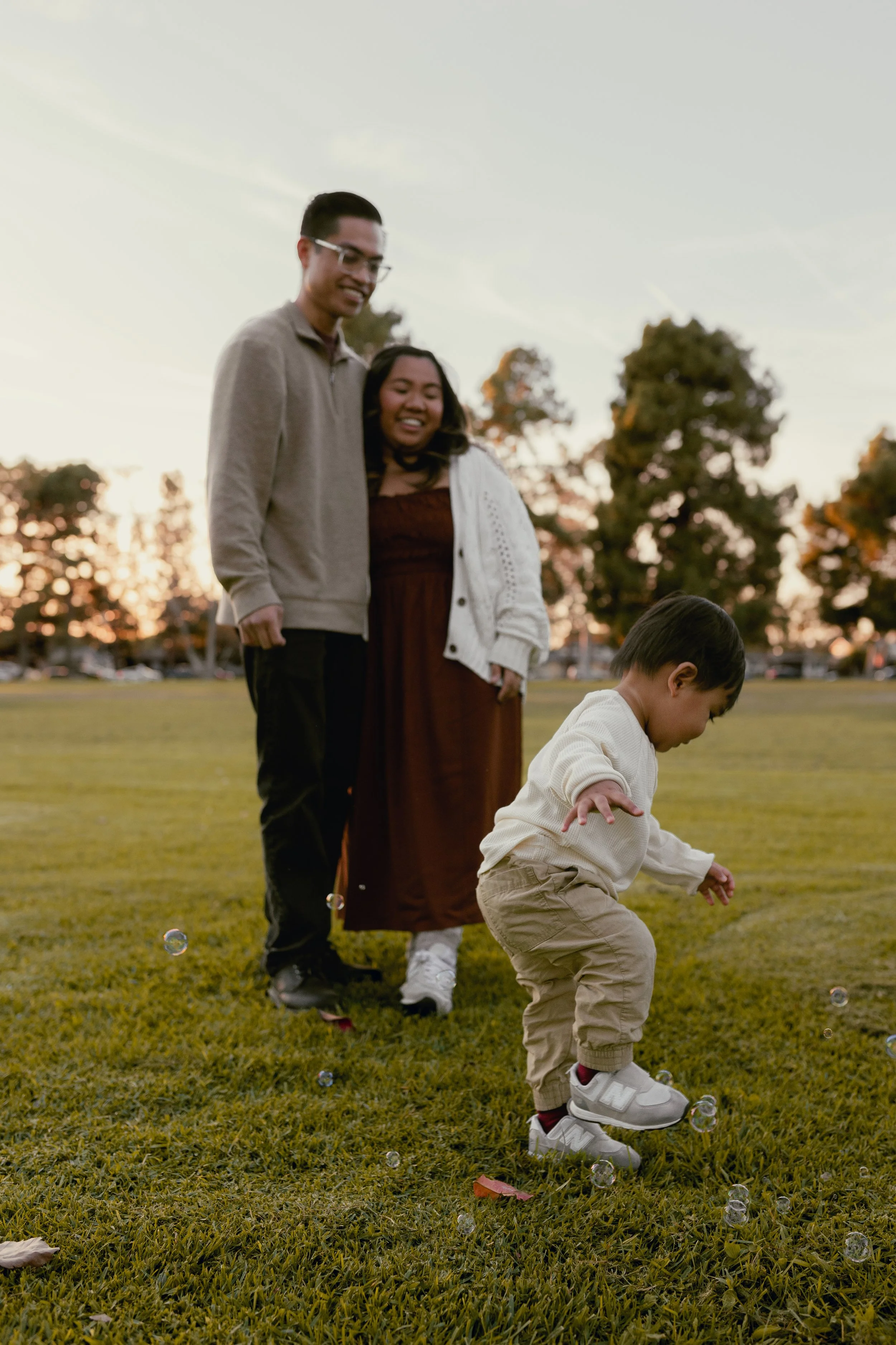 A young boy playing on the grass with bubbles while a man and woman watch and smile in a park during sunset.