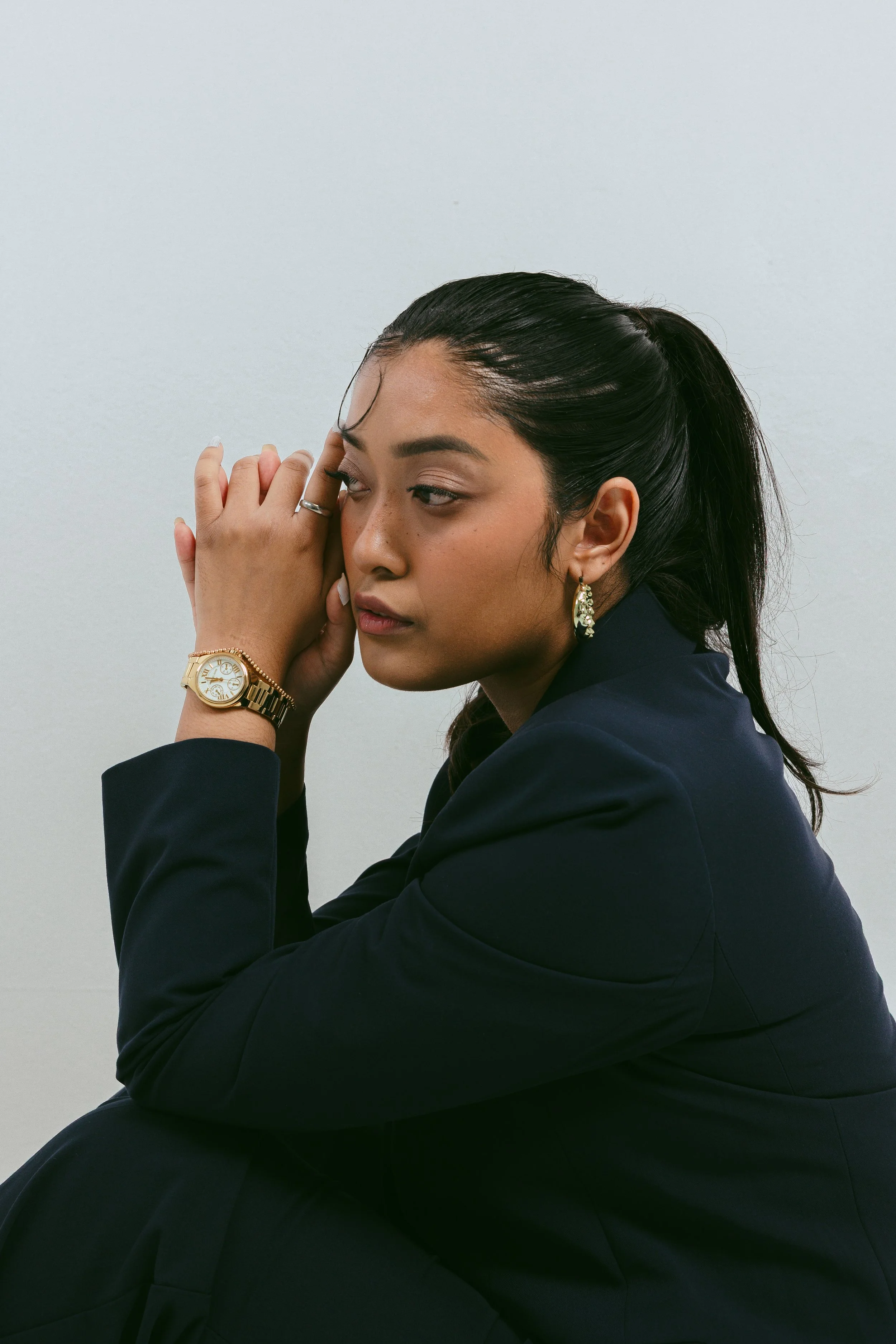 A woman with long dark hair styled in a ponytail, wearing makeup, earrings, a gold watch, a ring, and a navy blazer. She is resting her face on her hand with her eyes closed against a plain light gray background.